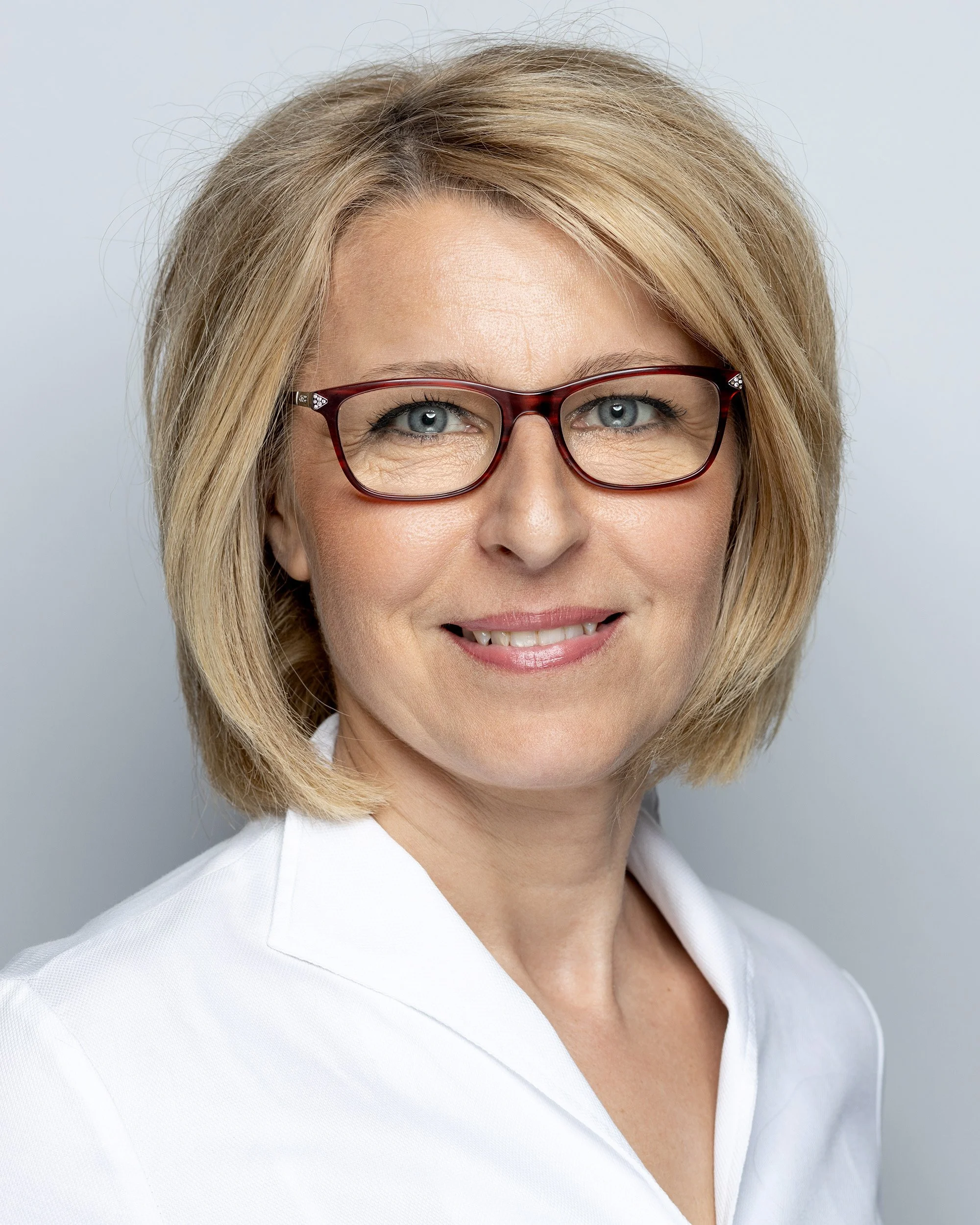 Close-up of a woman with blonde hair wearing glasses and a white blazer, smiling gently against a plain light background.