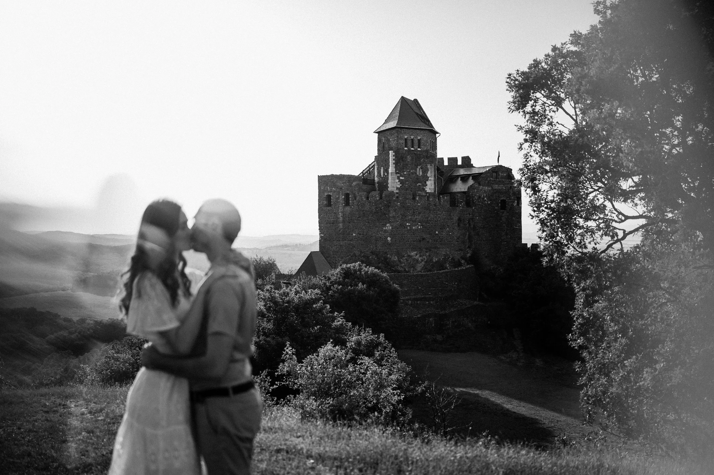 A black and white photo showing a blurred couple kissing in the foreground with a castle on a hill in the background surrounded by trees and open landscape.