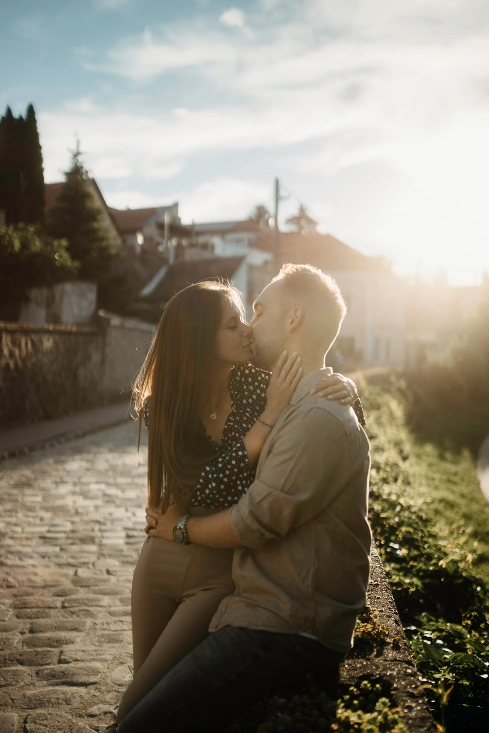 A couple sharing a kiss outdoors in warm sunlight during sunset, with a cobblestone street and houses in the background.