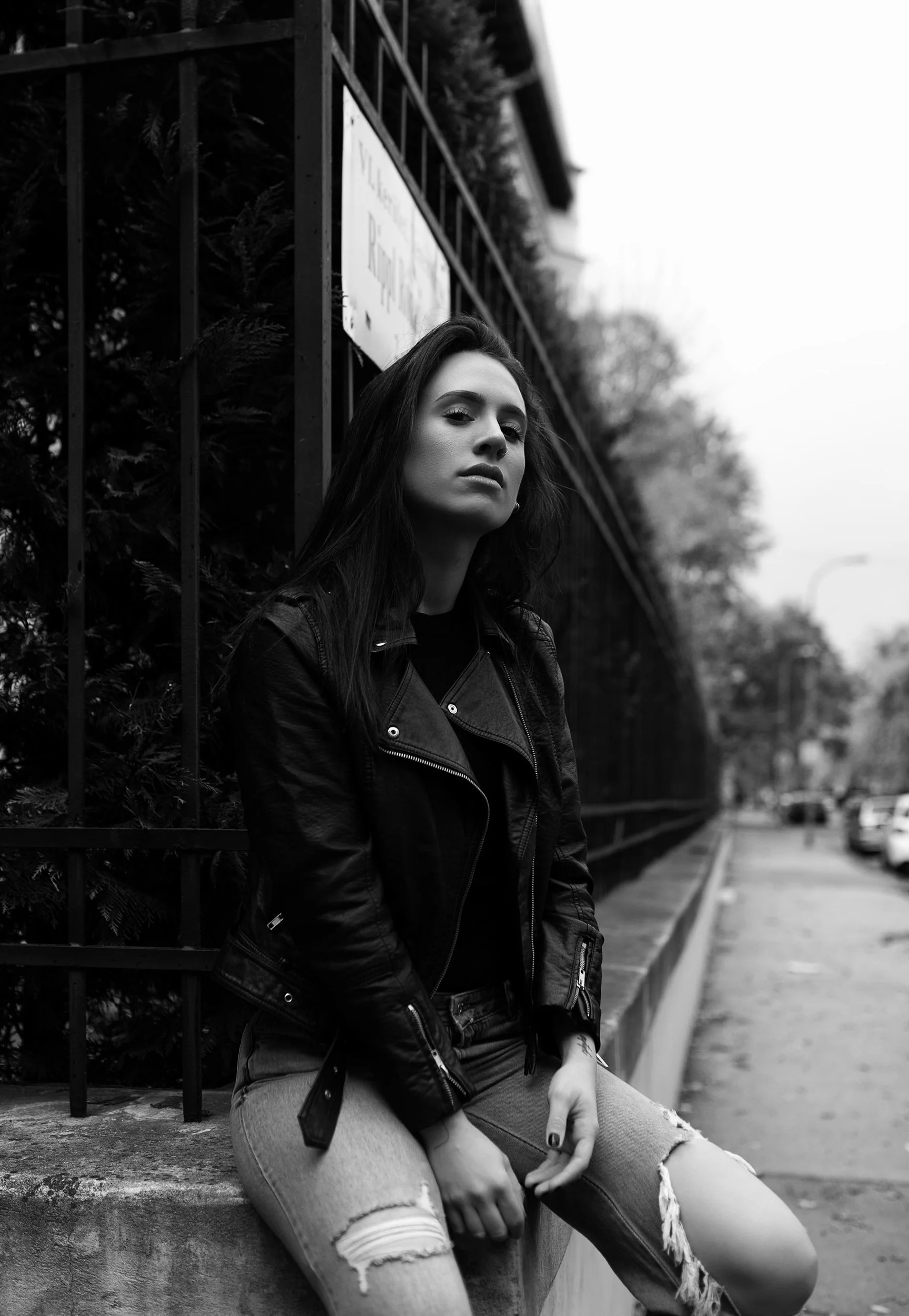 Black and white photo of a woman with long dark hair sitting on a concrete ledge, wearing a leather jacket and ripped jeans, with a serious expression, in an urban outdoor setting.