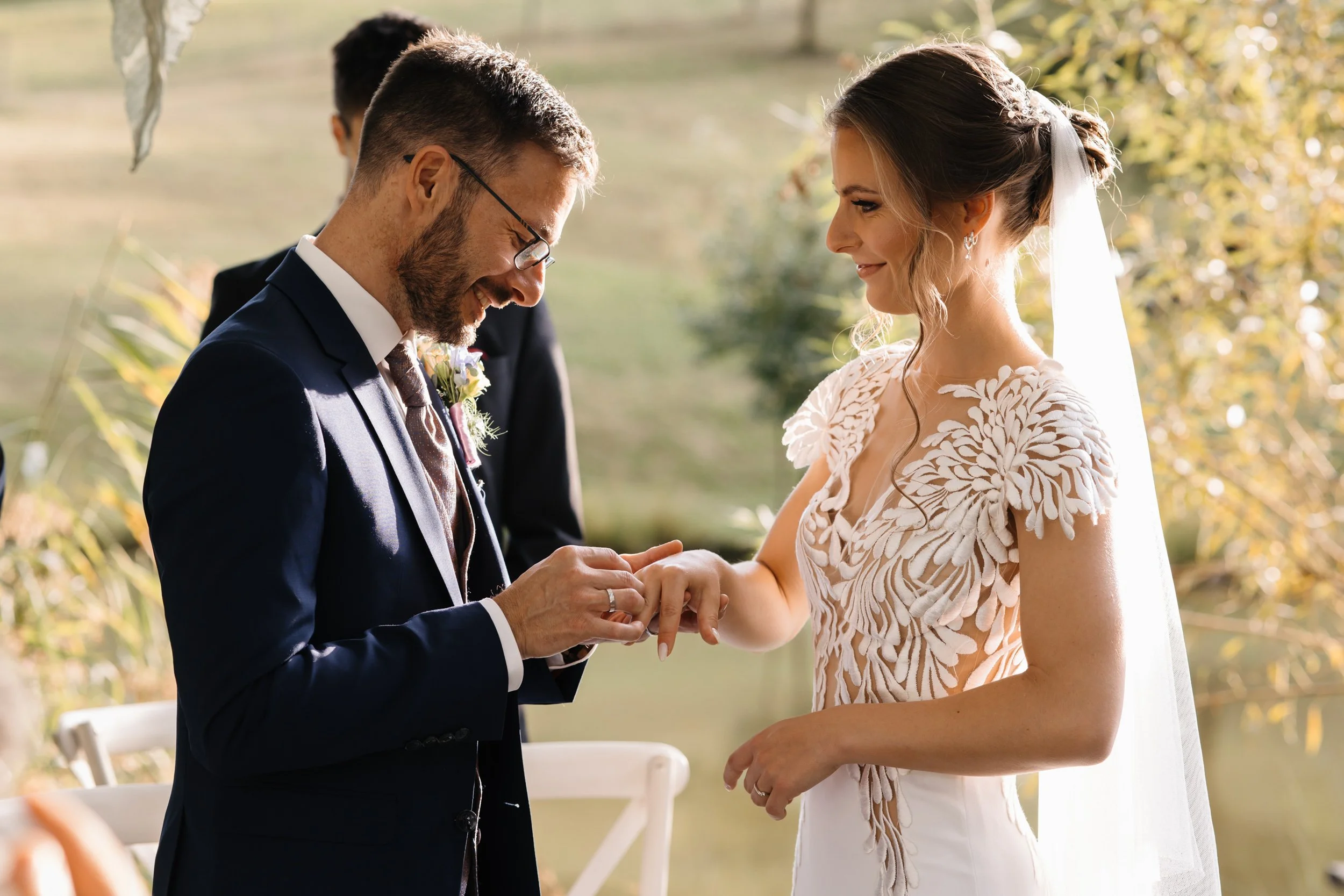 A bride and groom holding hands during their outdoor wedding ceremony, exchanging rings, with a man in the background.