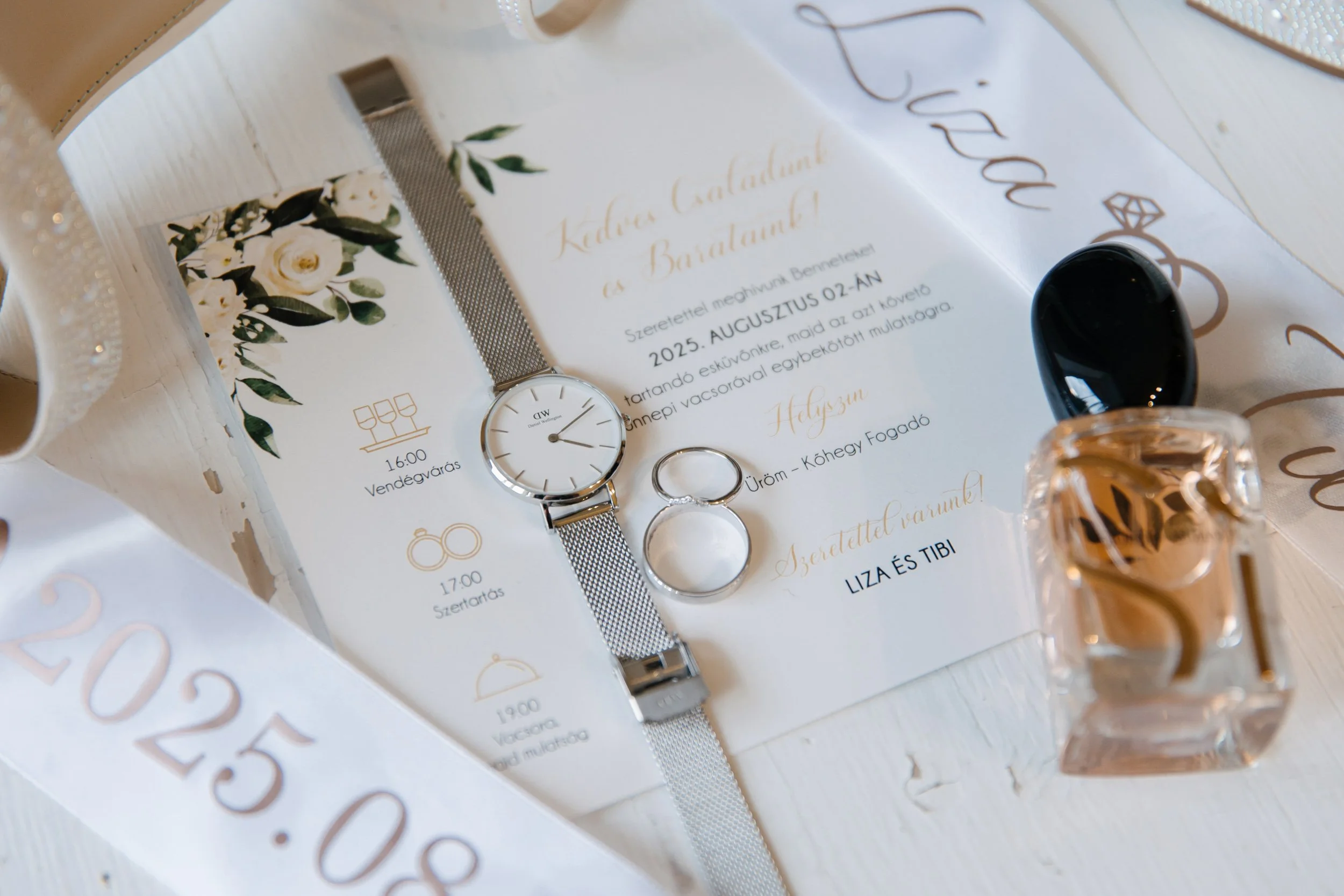 Wedding invitation with a floral design, a wristwatch, two rings, and perfume bottles on a white tablecloth.