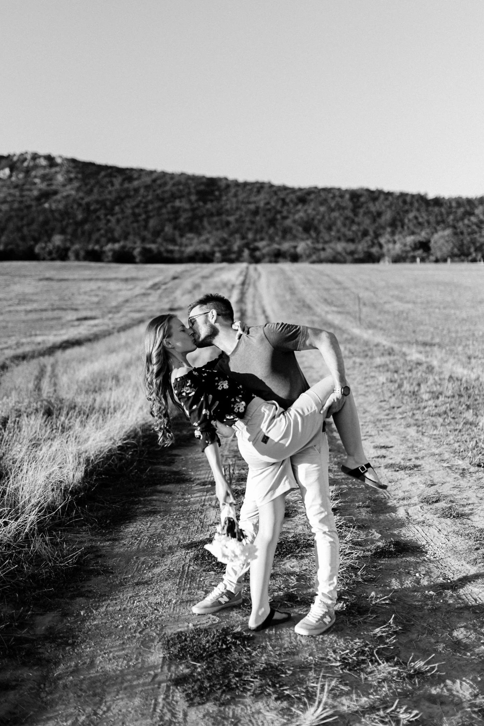 A black and white photo of a man lifting a woman in a field, with forested hills in the background, sharing a romantic moment.