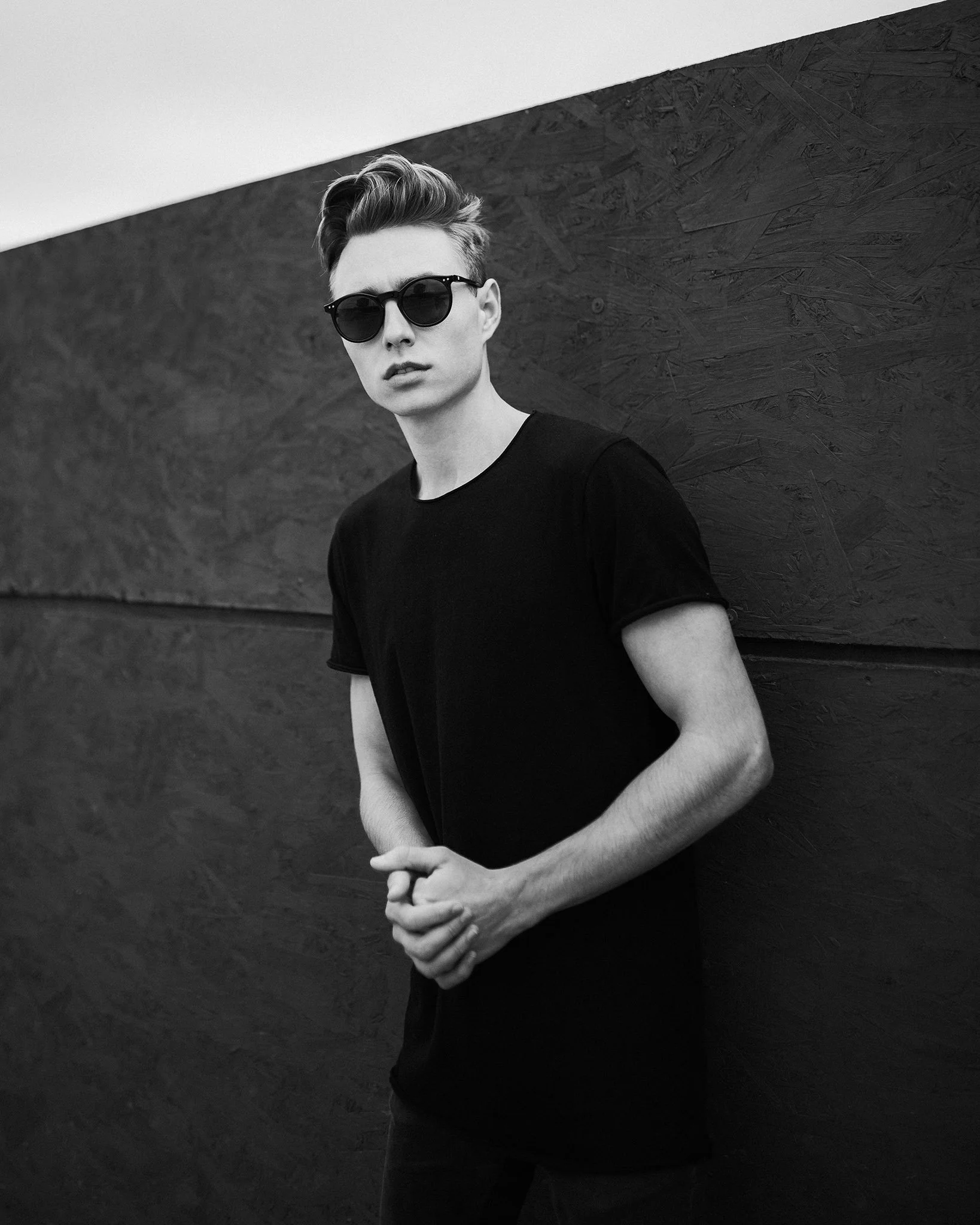 A young man with styled hair and wearing sunglasses and a black t-shirt, standing against a textured dark wall, looking directly at the camera with a serious expression.