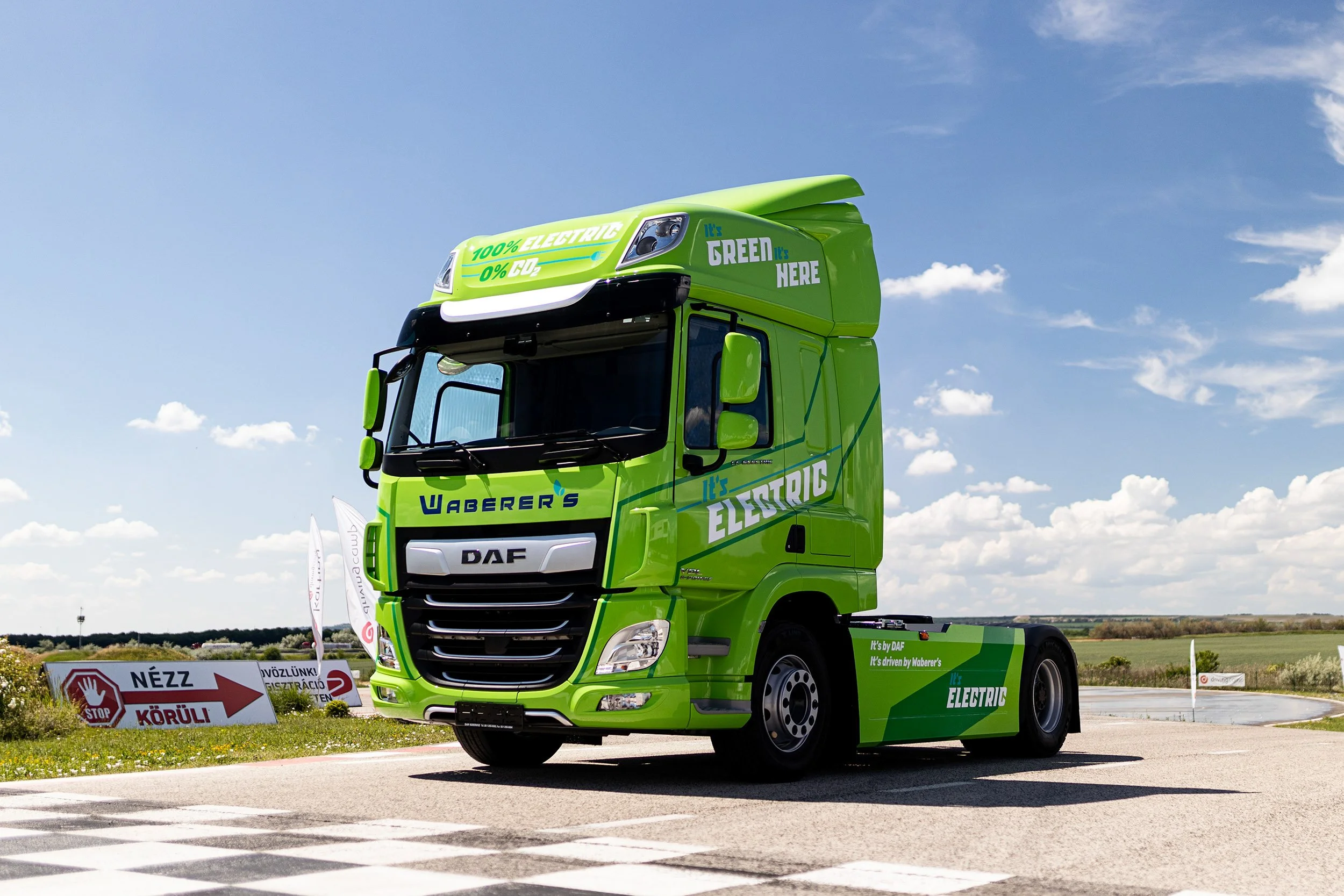 Green electric truck with branding promoting eco-friendly transportation, parked outdoors under a blue sky with clouds, near road signs indicating a stop and caution in multiple languages.