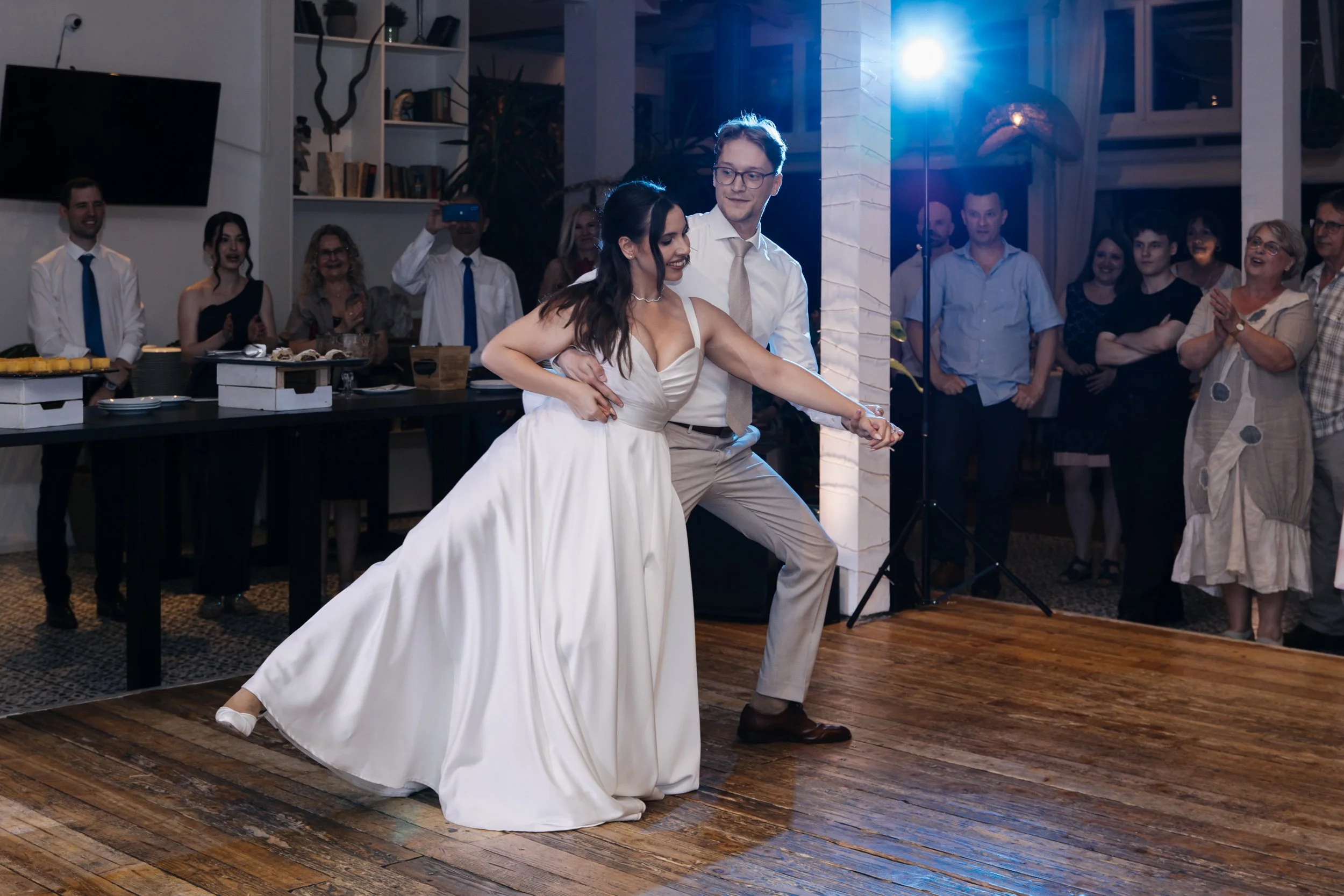 A bride and groom dancing at their wedding reception, surrounded by guests in a decorated indoor venue with wooden floors and bookshelves.