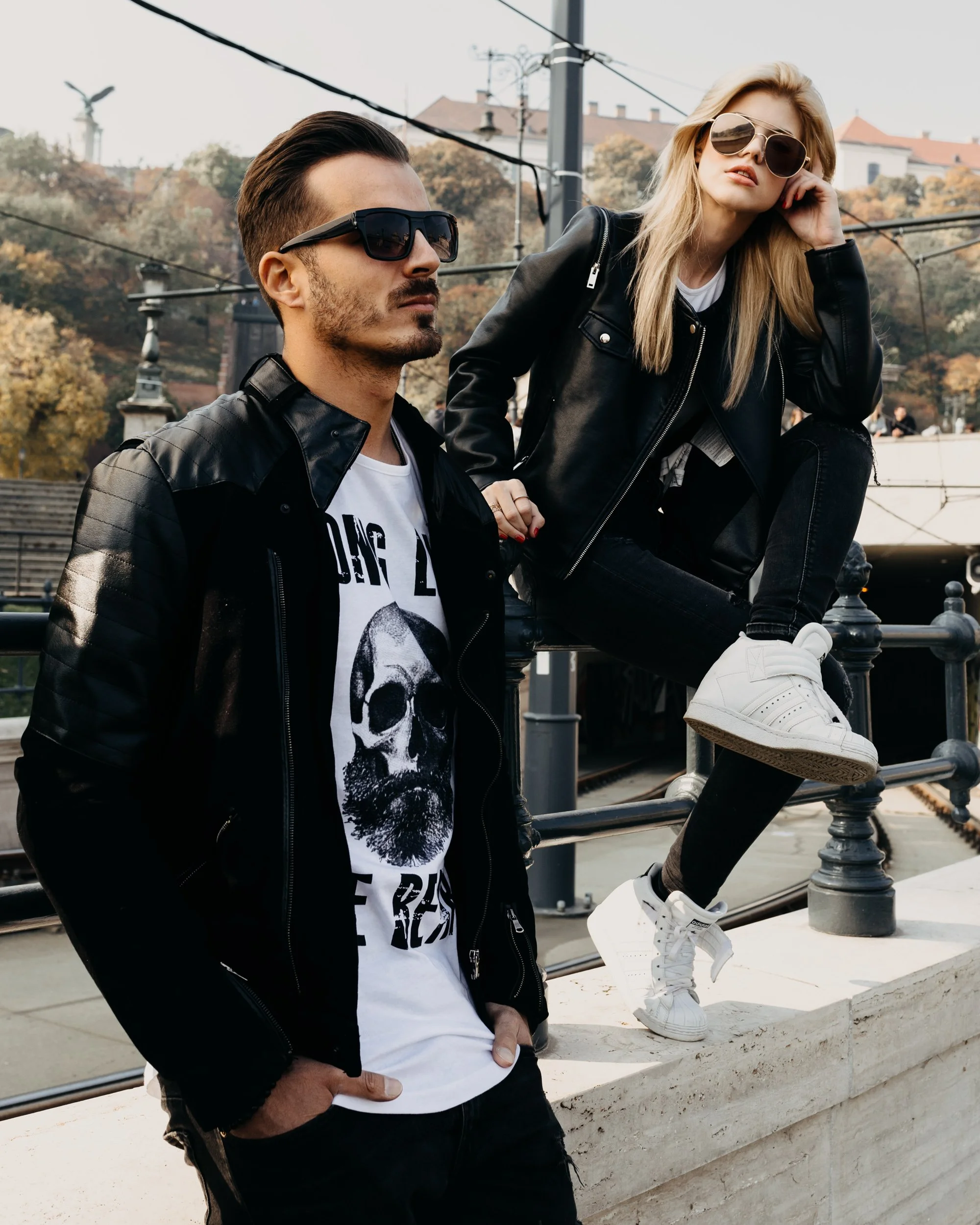 A man and a woman in sunglasses, dressed in black leather jackets, posed outdoors with autumn foliage in the background. The woman is sitting on a railing with her leg raised, wearing white sneakers, while the man is standing with hands in his pocket