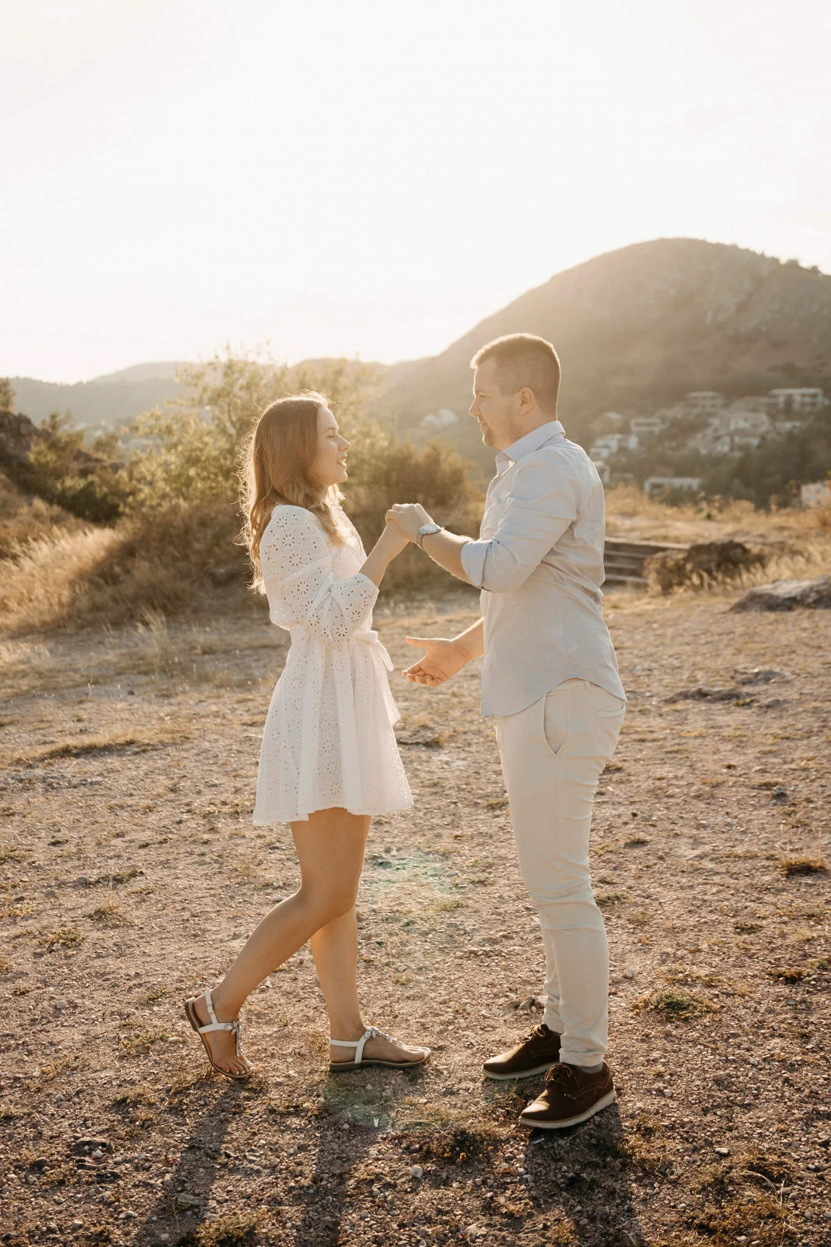 A couple holding hands and dancing outdoors during sunset, with mountains and a few houses in the background.