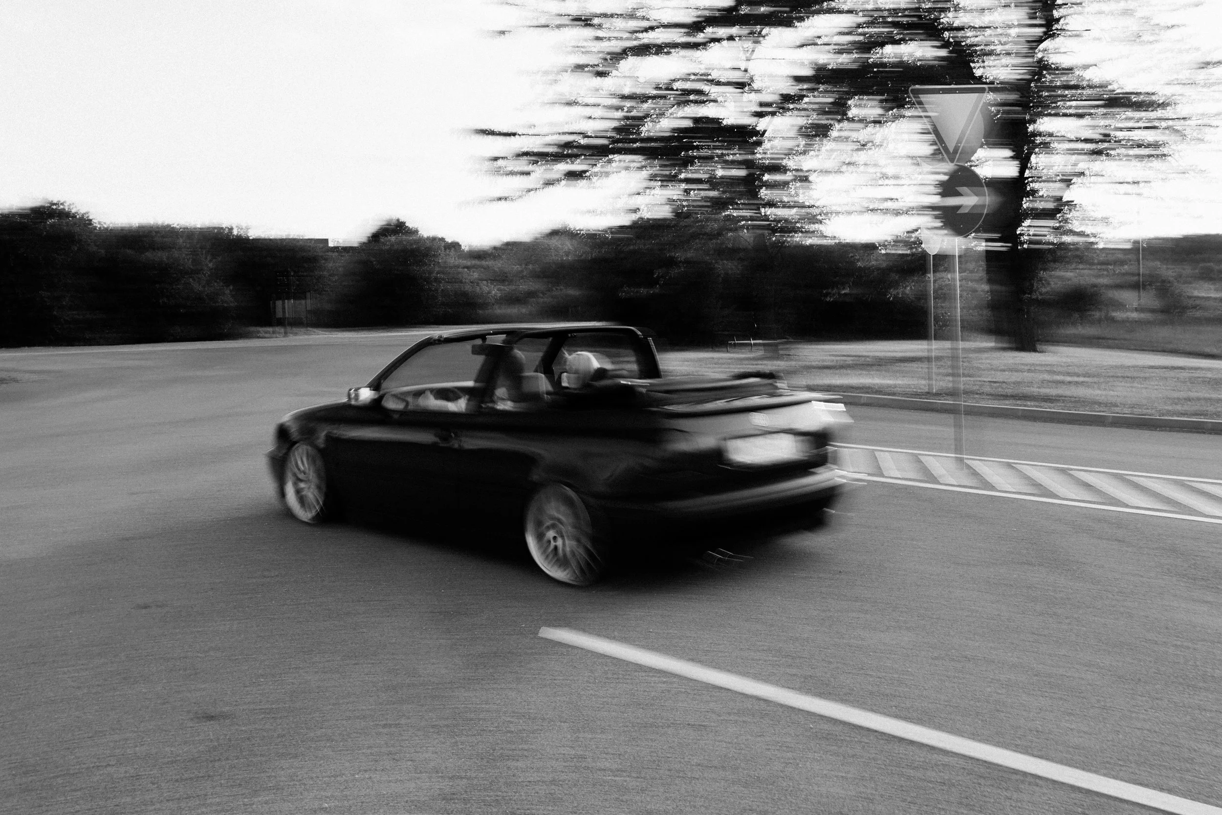 A black convertible car driving on a curved road with trees and a traffic sign in the background, captured in motion blur in black and white.