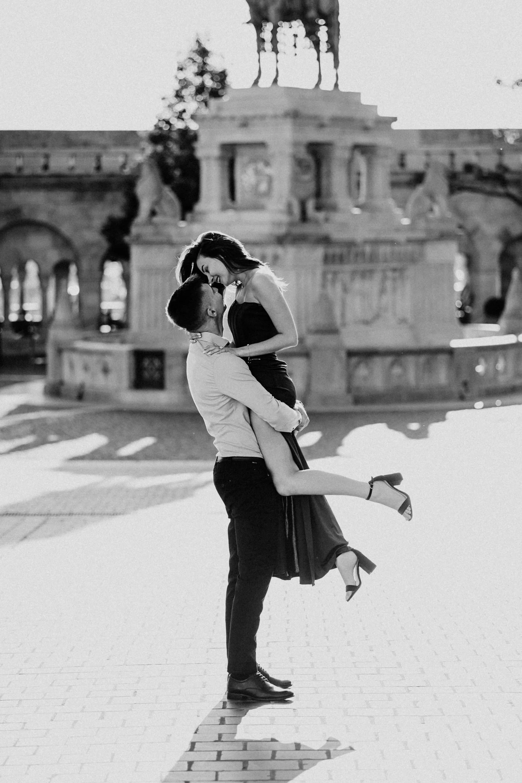 A black and white photo of a couple dancing outdoors, with the man lifting the woman in his arms. The woman is smiling and looking at the man, who is also smiling. There is a large elaborate building in the background, with statues and architectural 
