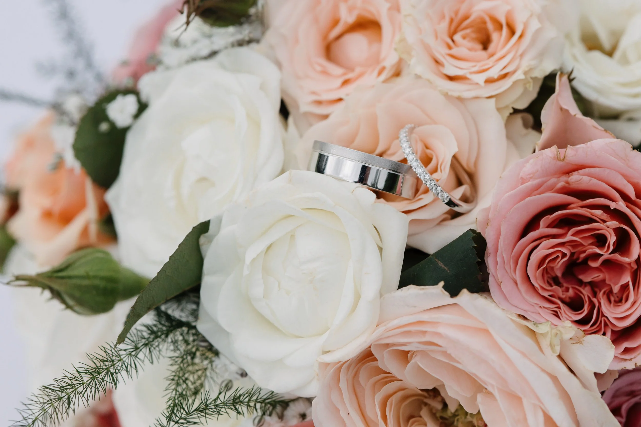 Close-up of a wedding bouquet with pink and white roses, green leaves, and two wedding rings resting on the flowers.