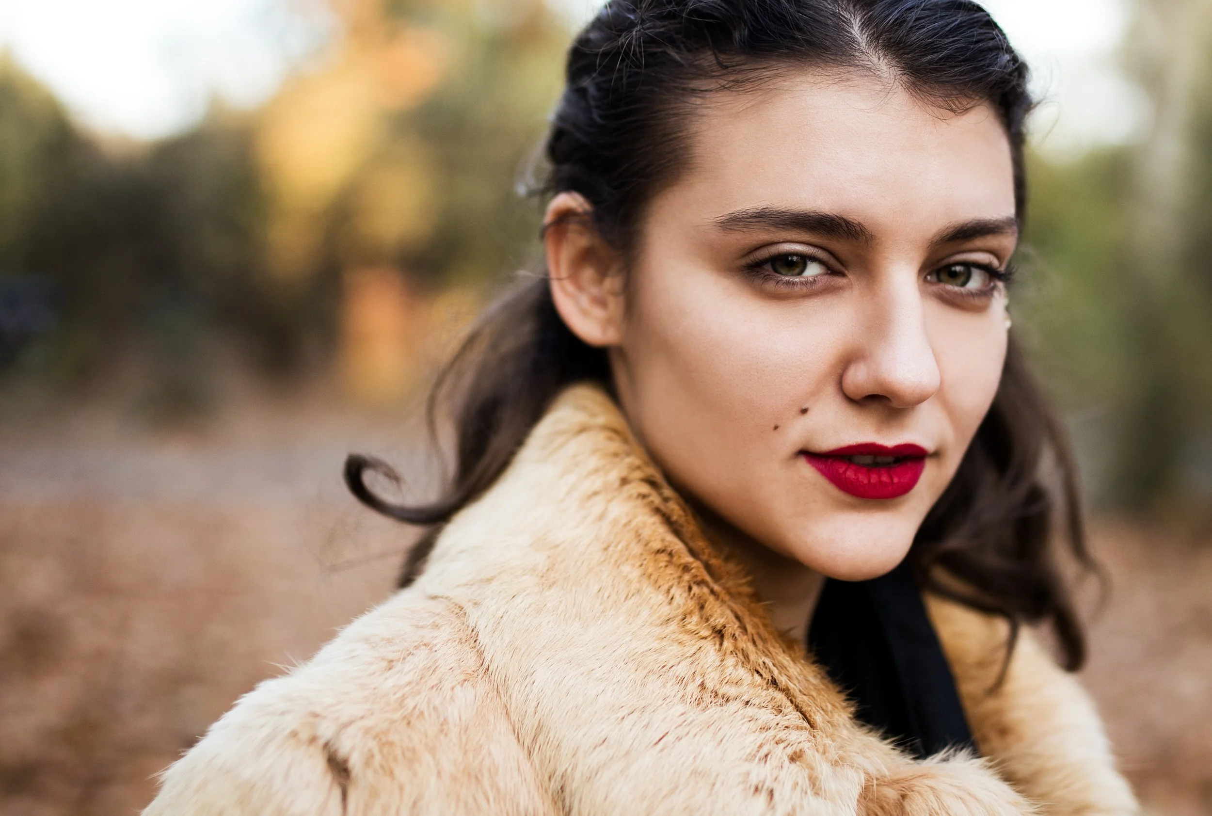 Close-up of a woman with dark hair, red lipstick, and a tan fur coat outdoors with autumn foliage in the background.