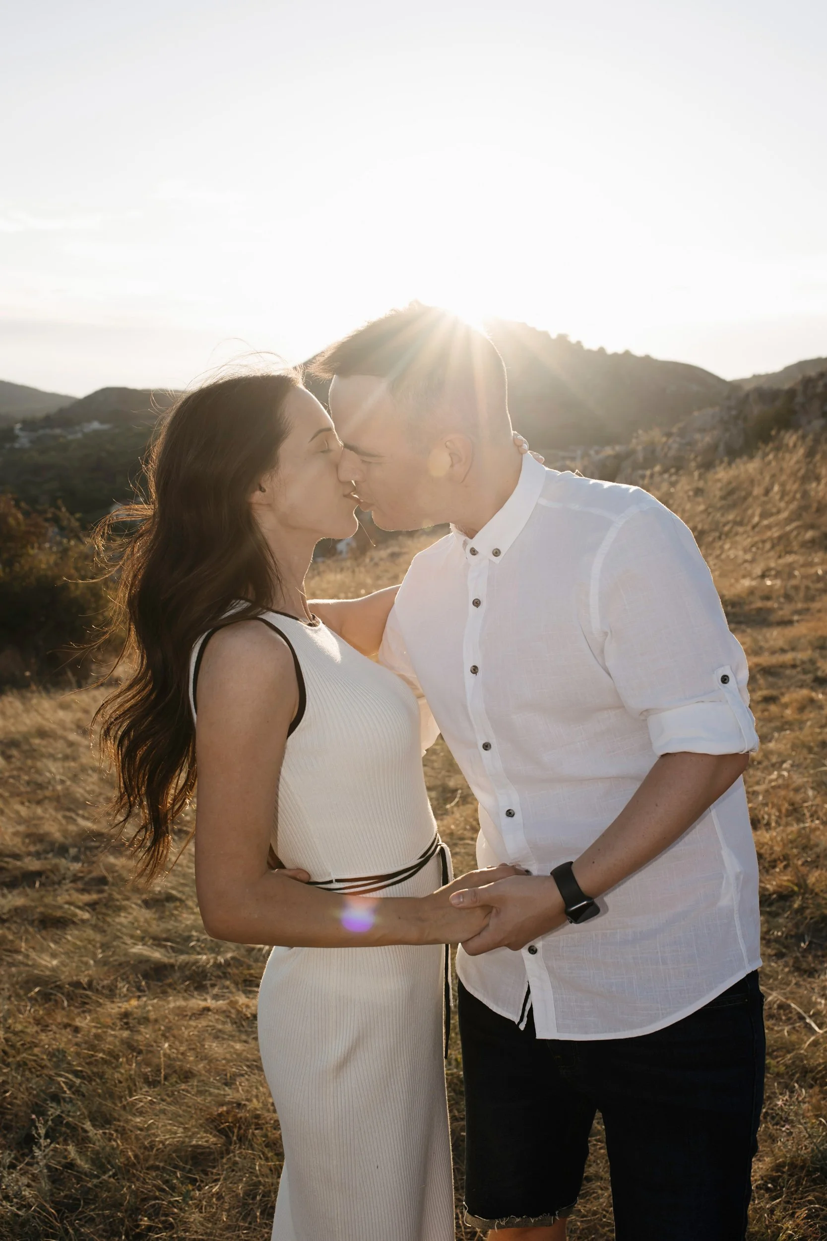A couple standing outdoors during sunset, about to kiss, with rolling hills in the background.