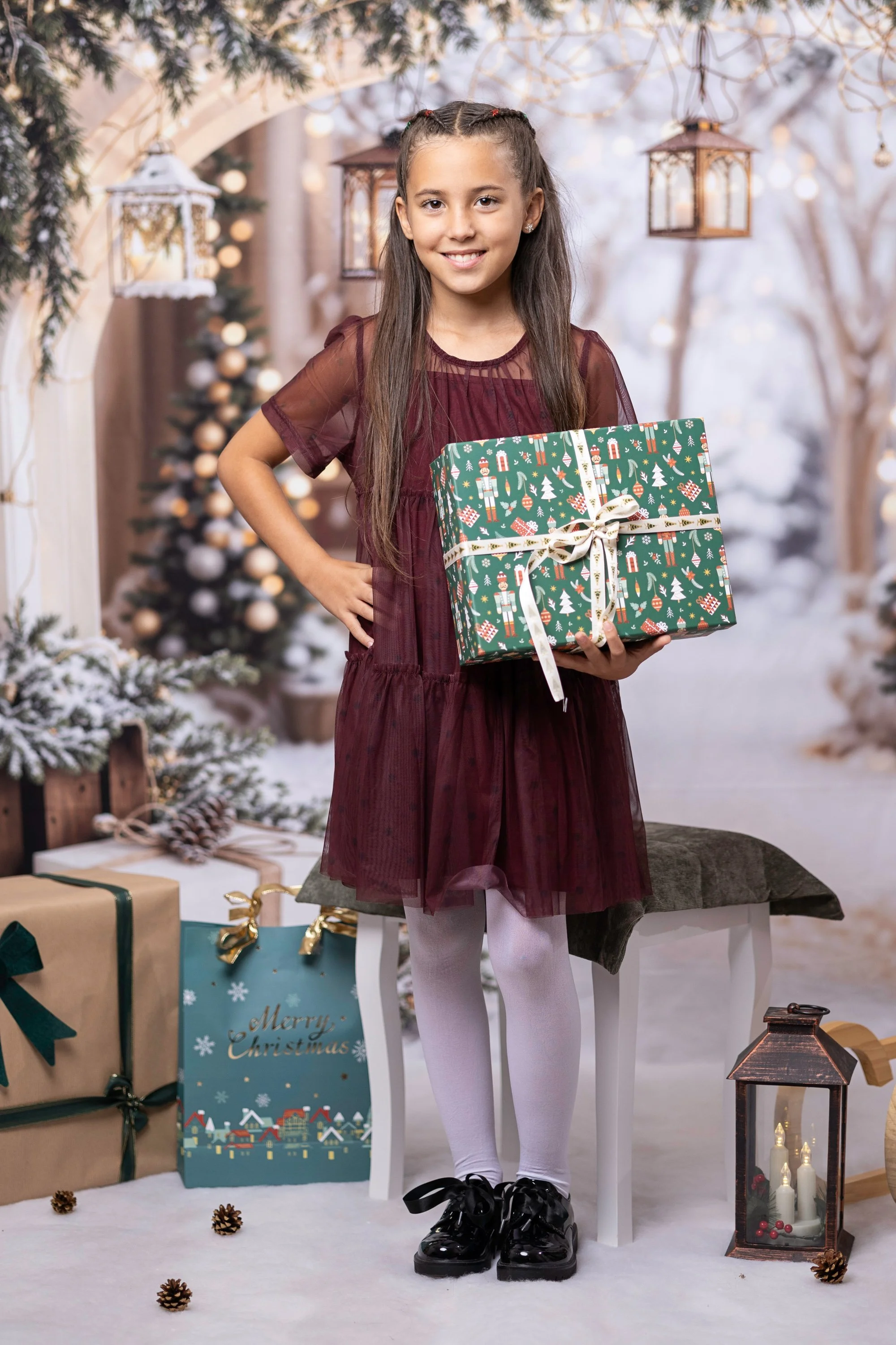 A young girl with long brown hair standing in front of a festive Christmas scene, holding a wrapped present. She is wearing a burgundy dress, white tights, and black shoes. The background features Christmas trees, decorated packages, and hanging lant