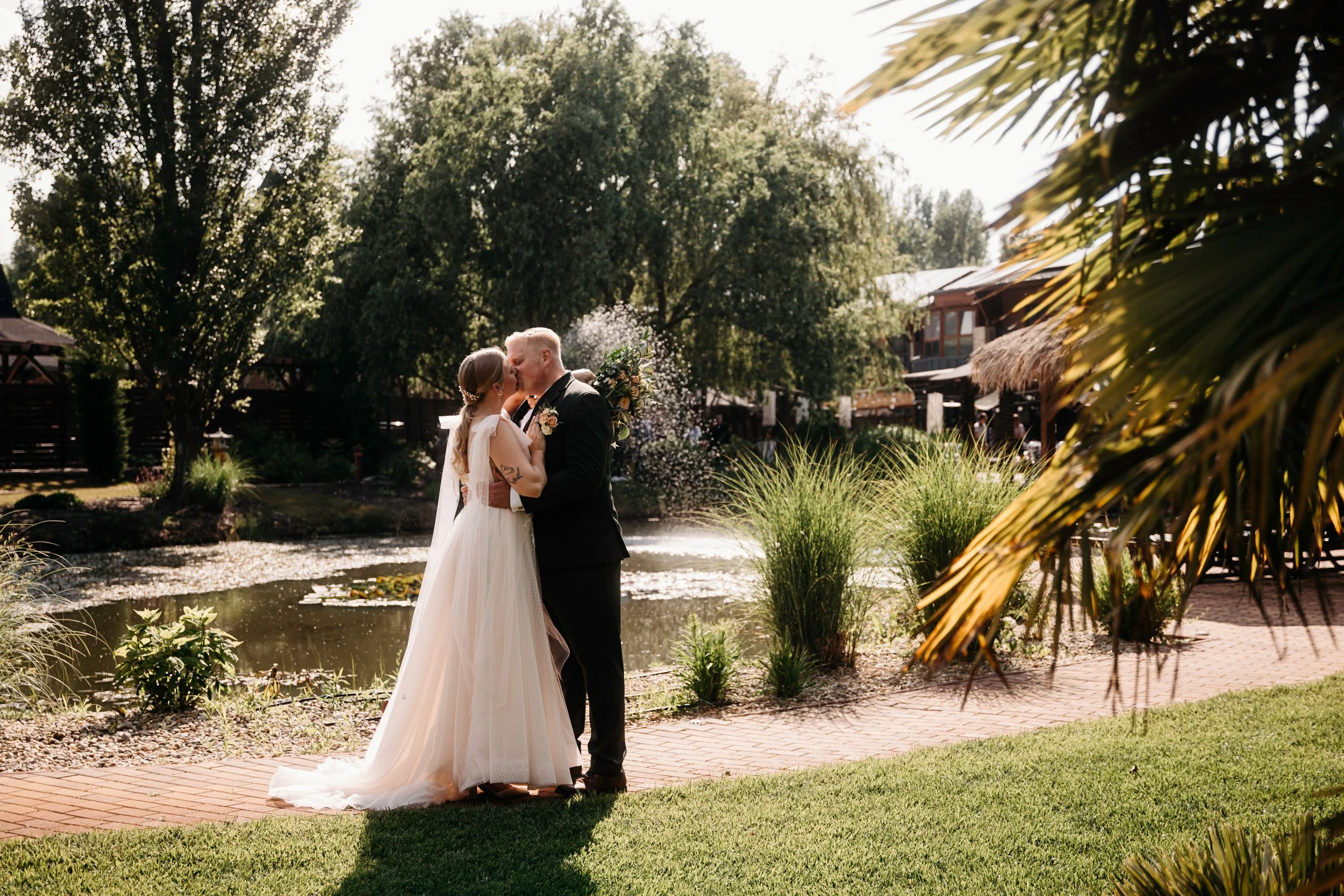 A bride and groom embrace and kiss by a pond surrounded by lush greenery and trees, with a building in the background on a sunny day.