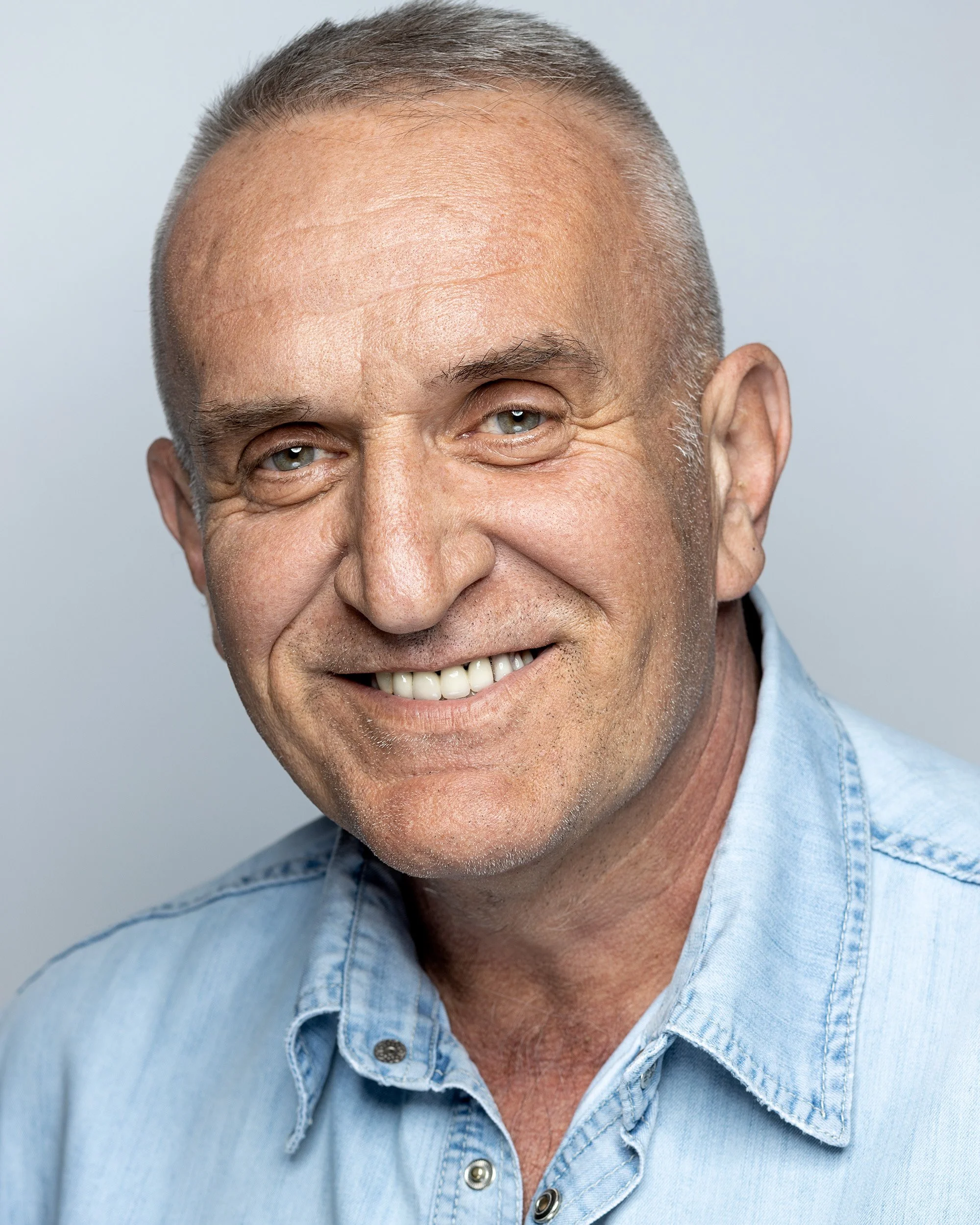 Close-up photo of an elderly man smiling, showing his teeth, with gray hair, partly visible face, and wearing a light denim shirt against a plain light gray background.