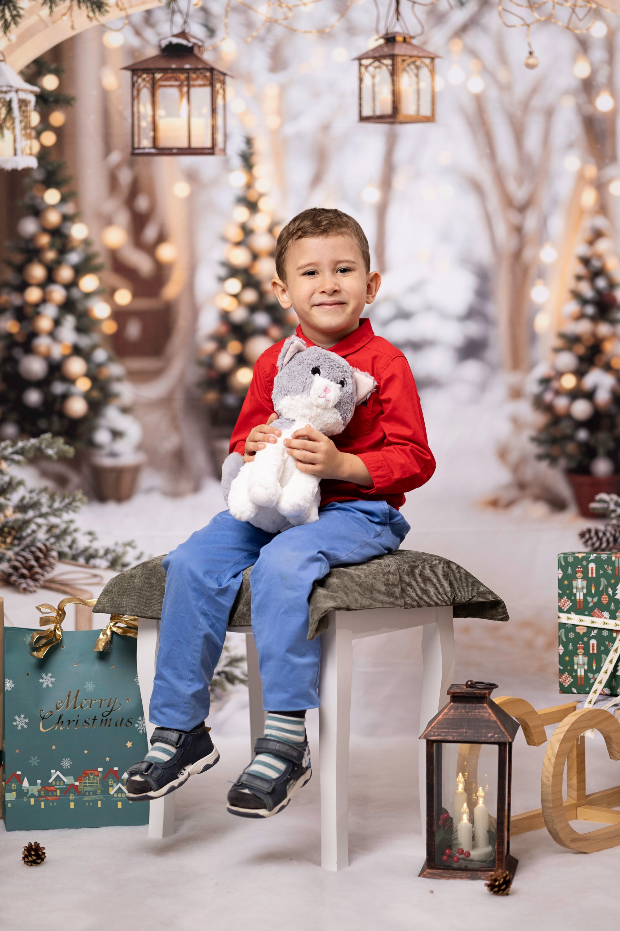 A young boy sitting on a white stool outdoors in a winter scene, holding a gray stuffed animal. The background features snow-covered trees, decorated Christmas trees, hanging lanterns, and Christmas gifts, with a festive and cozy holiday atmosphere.