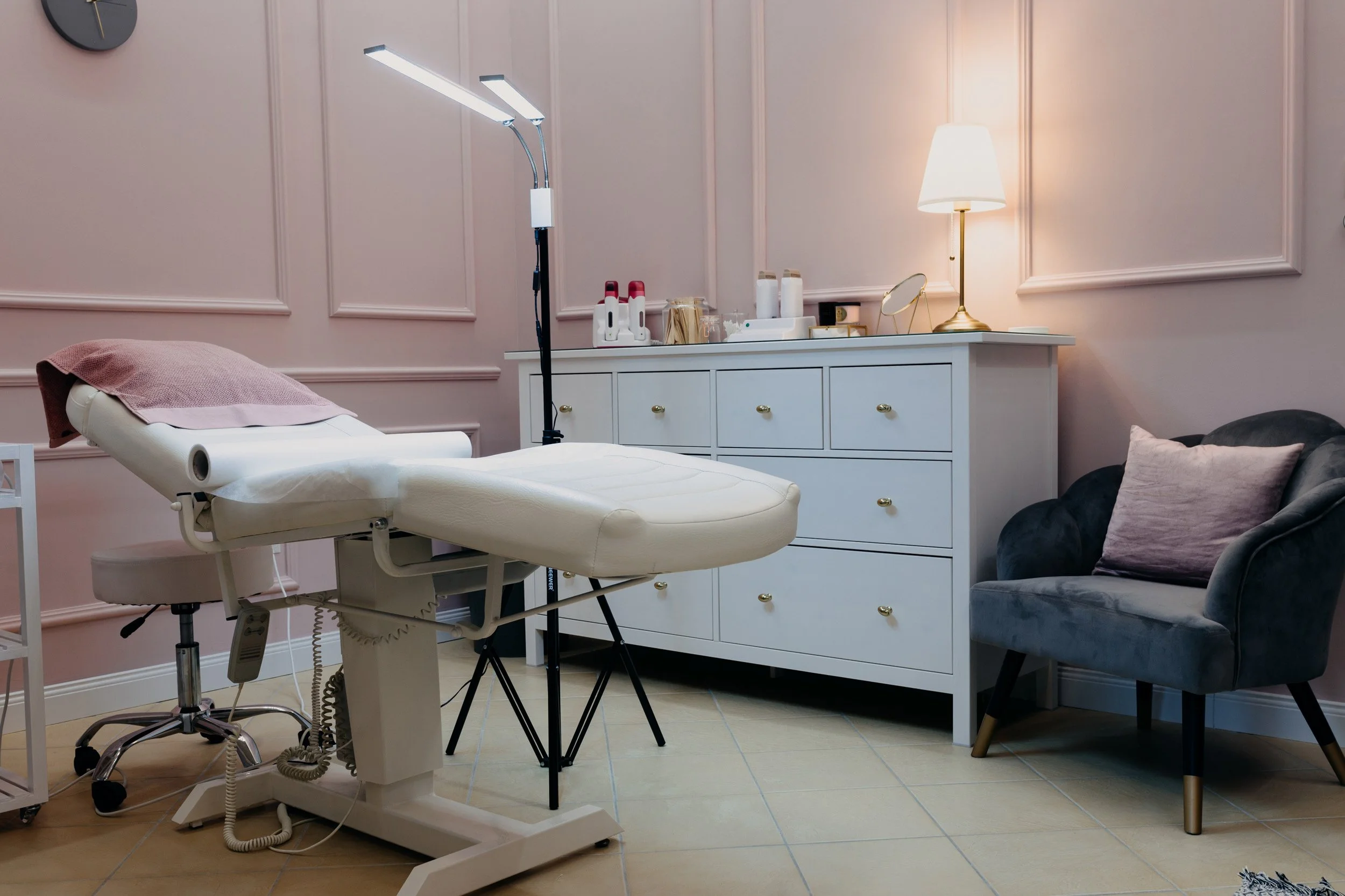 A treatment room with a reclining medical chair, a white cabinet with medical supplies, a small round mirror, a table lamp, a gray armchair with a pink pillow, and a wall clock.