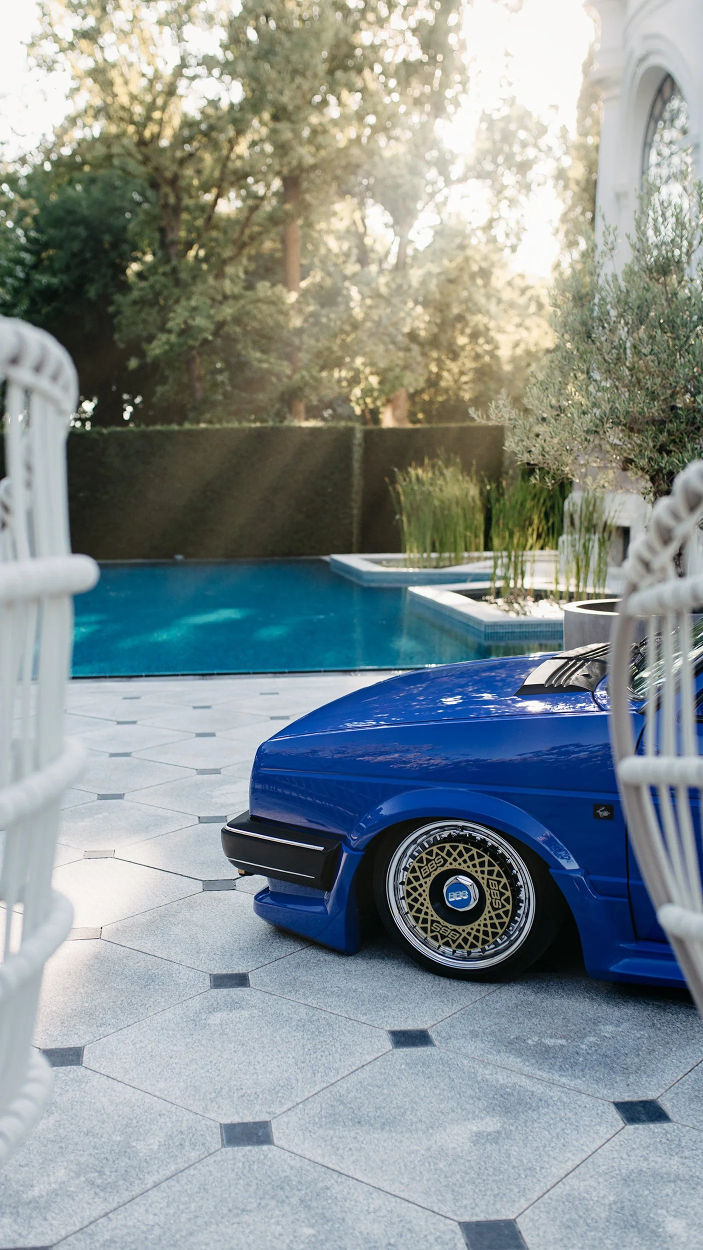 A blue car parked by a swimming pool in a backyard, with white patio furniture and green plants, under sunlight filtering through trees.