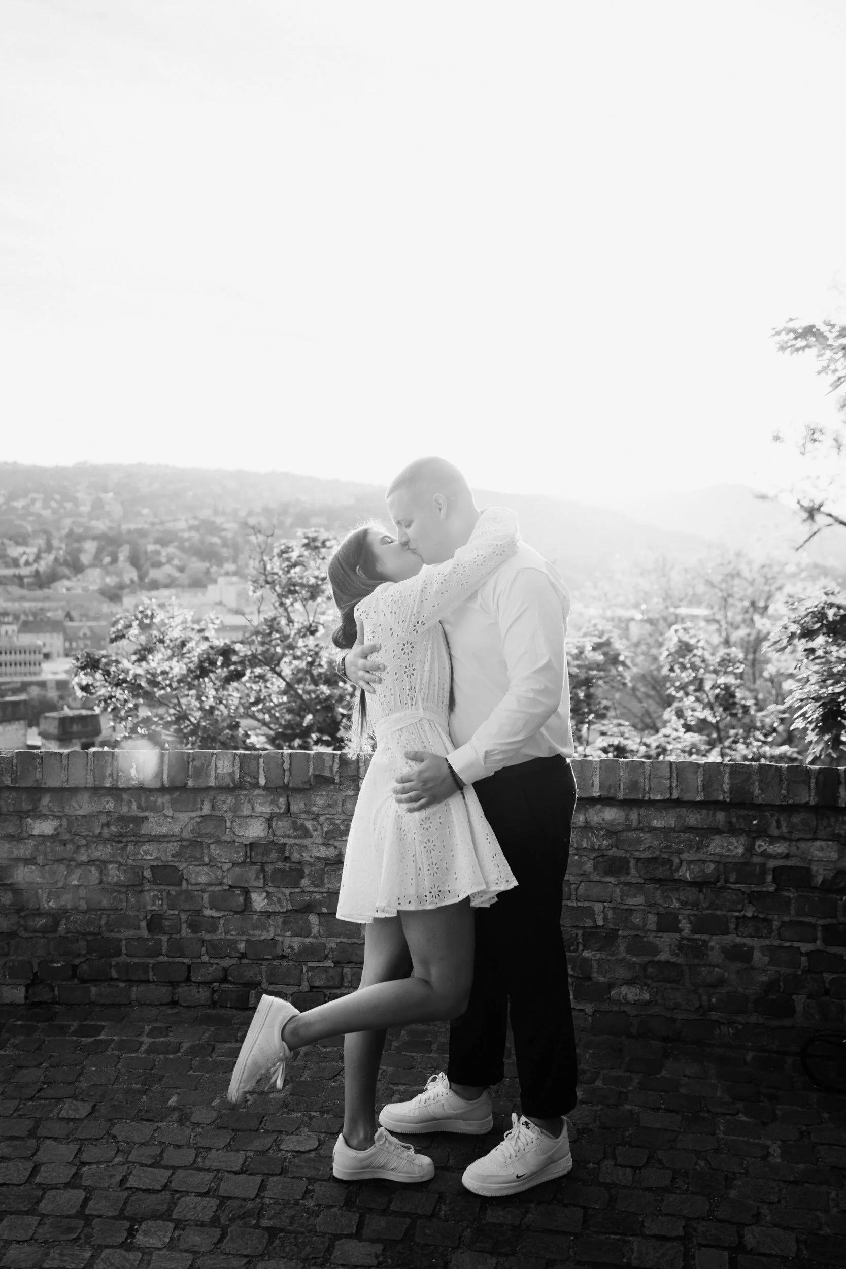 A black and white photo of a couple kissing on a rooftop with a city view in the background. The woman is wearing a dress and sneakers, and the man is dressed in a shirt, trousers, and sneakers. The woman has her legs slightly raised and wrapped arou