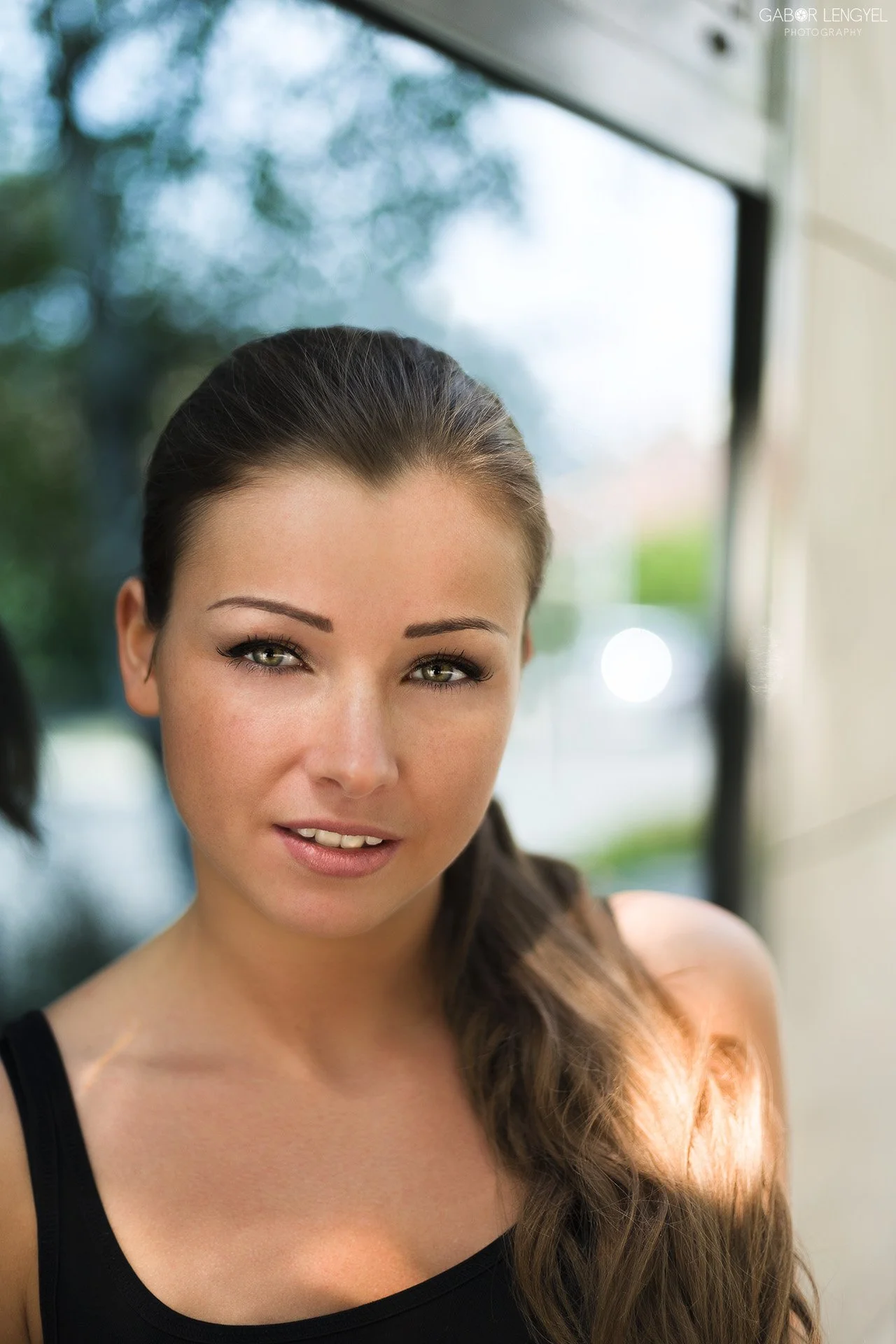 Close-up of a young woman with long, brown hair, fair skin, and hazel eyes, smiling softly, wearing a black tank top, standing near a window with blurred greenery outside.