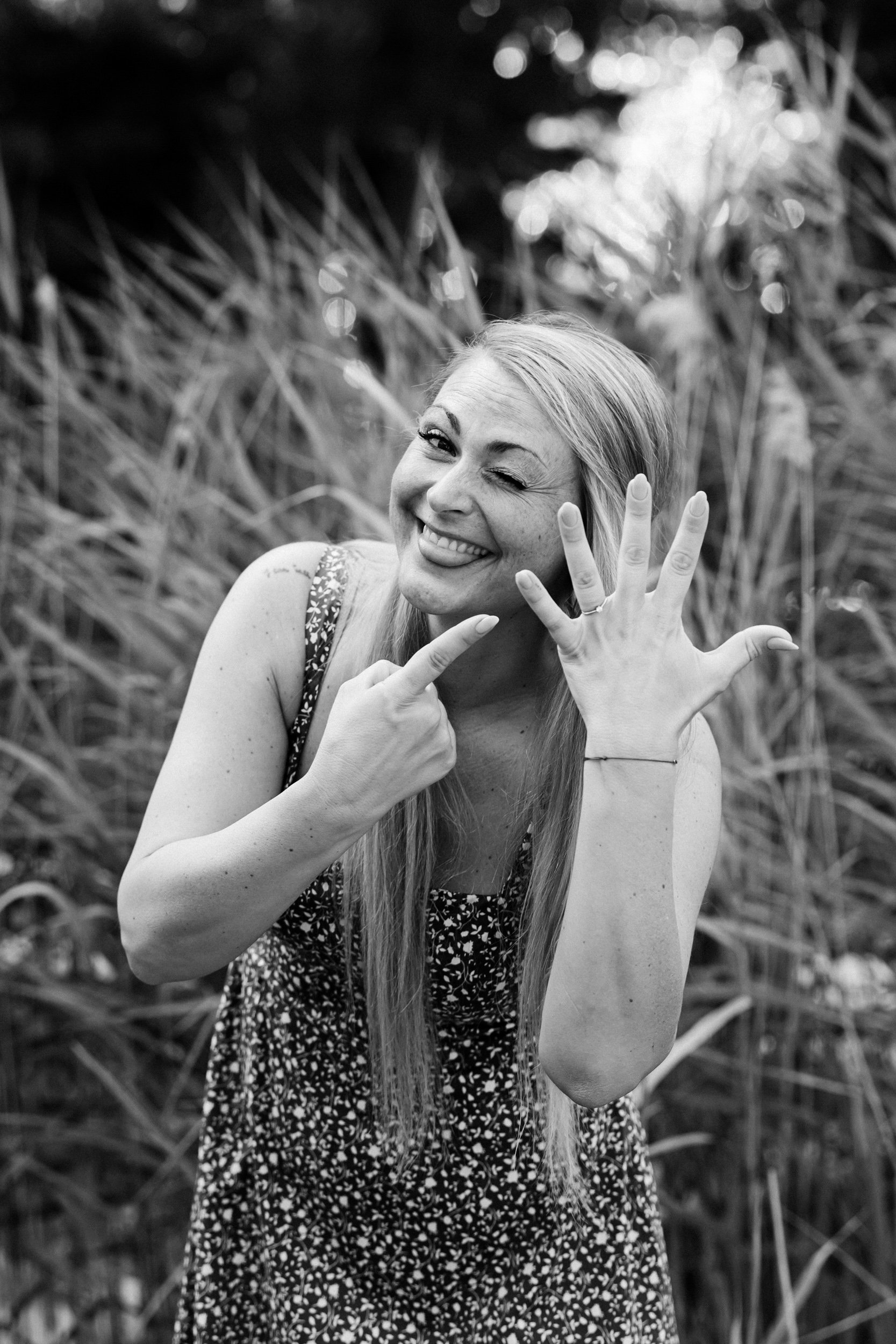 Black and white photo of a woman smiling and winking at the camera, making a playful gesture with her hand, in a natural outdoor setting.