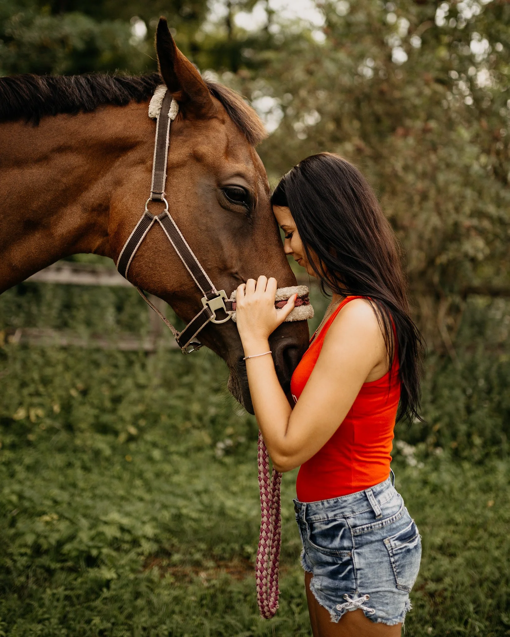 A young woman with long dark hair and a red tank top standing close to a brown horse, touching foreheads, outdoors with trees and greenery in the background.