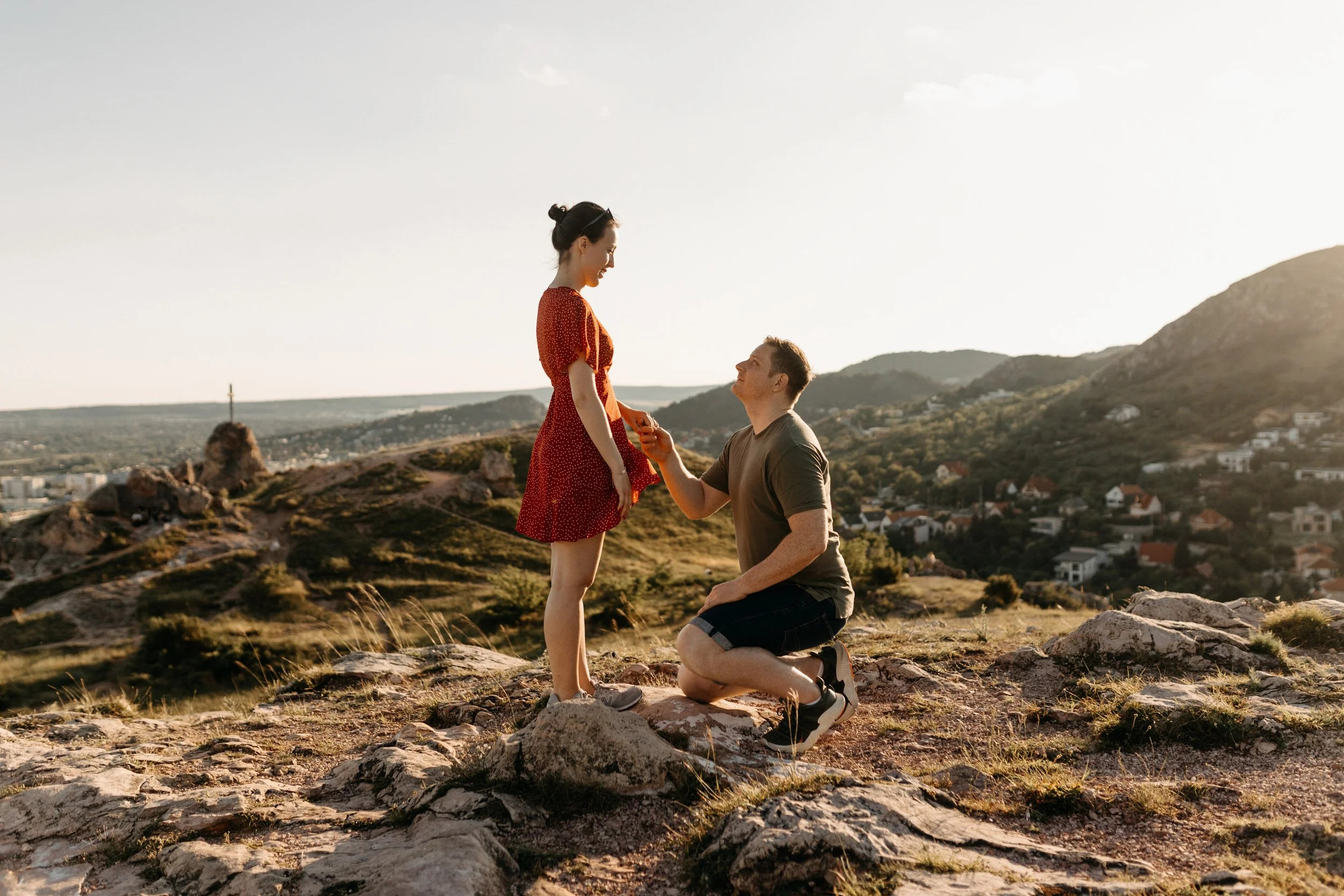 A man is kneeling on one knee on rocky terrain, proposing to a woman standing in front of him, with a scenic view of hills and a town in the background during sunset.