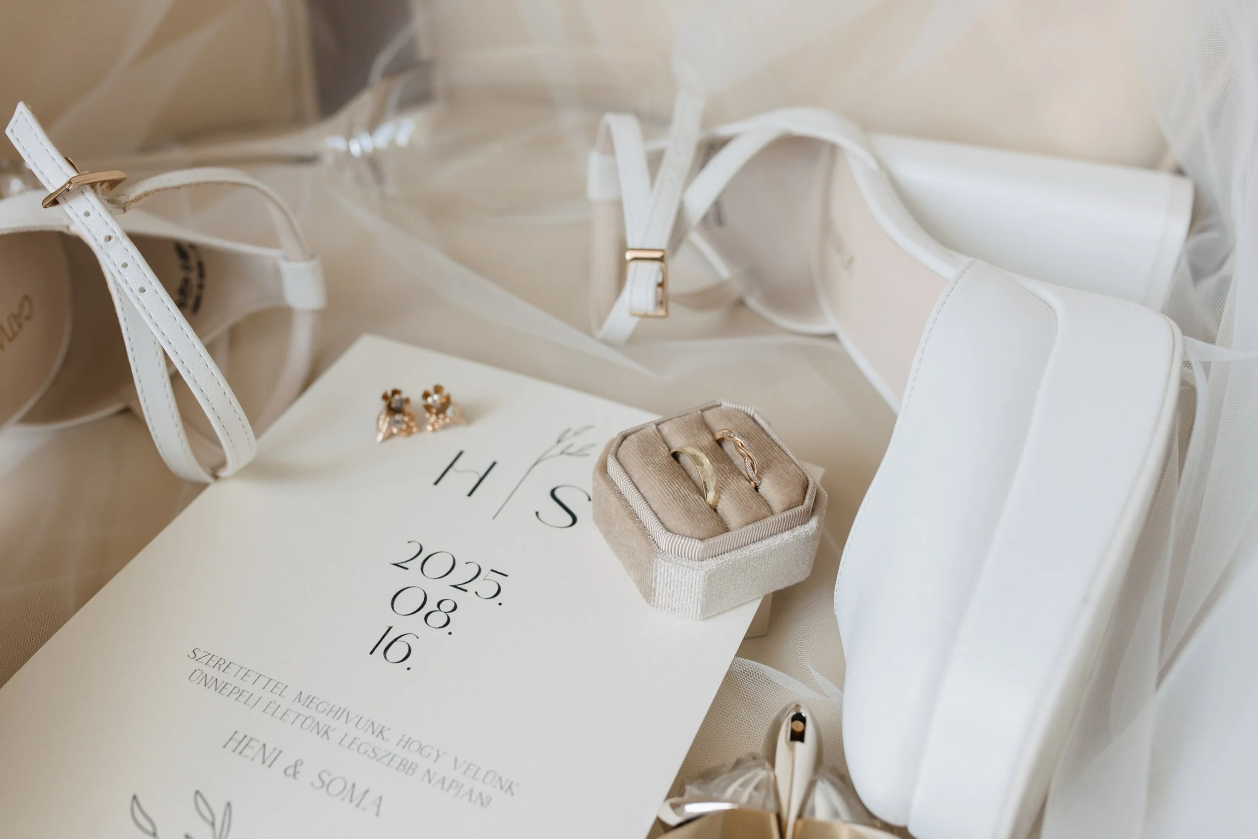 A table with wedding accessories including white sandals with ankle straps, a pair of earrings with floral designs, a small ring box with rings inside, and a wedding program or invitation with date and event details. The items are arranged on a white