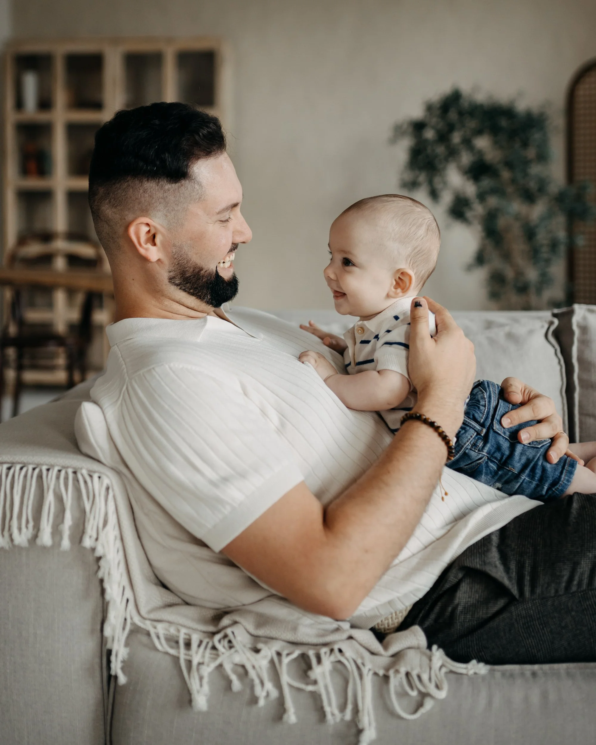 A man and a young child sitting on a cream-colored sofa. The man is smiling joyfully, holding the child cheek to cheek. The child is looking at the man with a happy expression. Behind them, a bookshelf and a green plant are visible in the background.