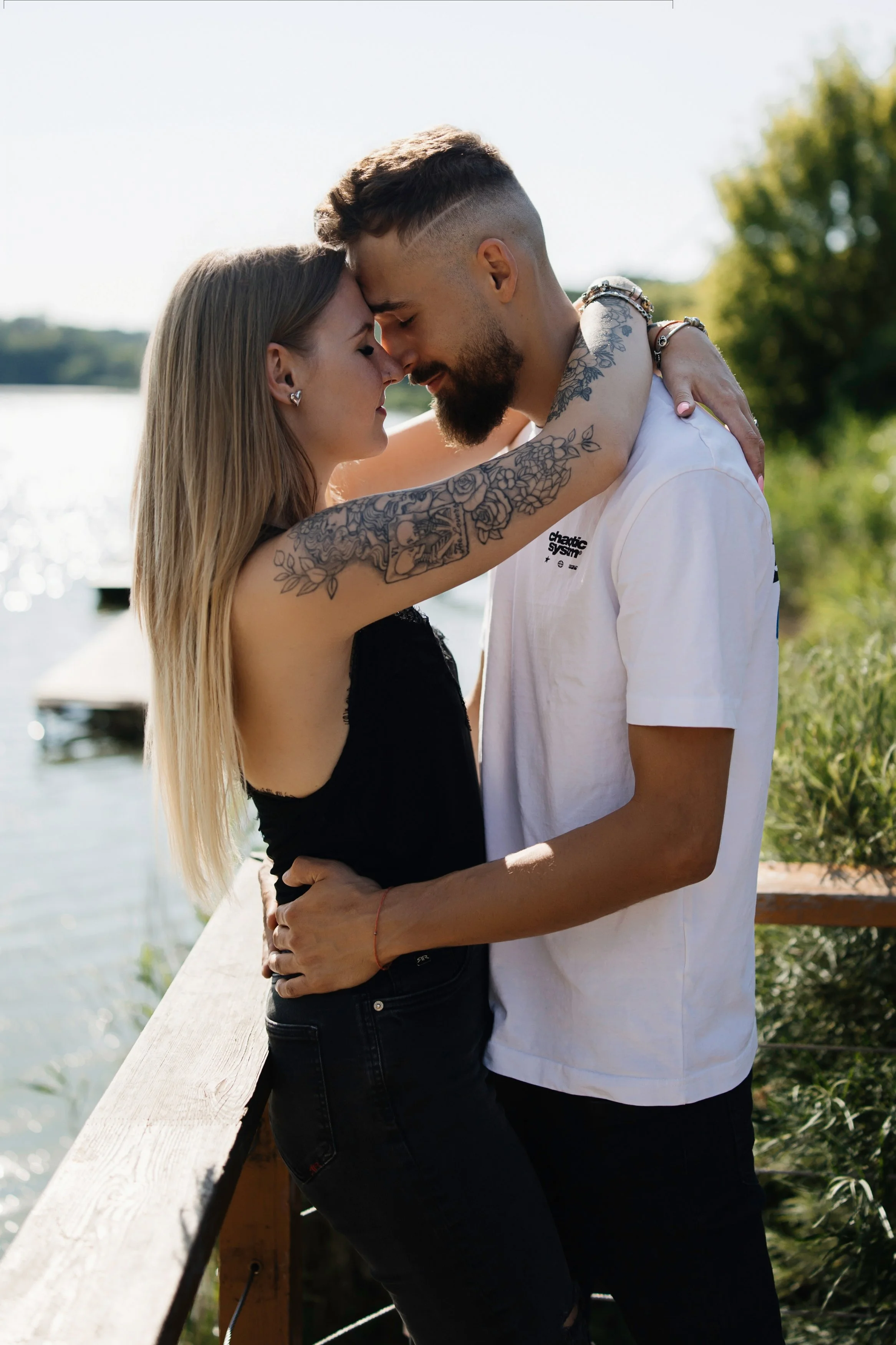 A couple with their foreheads touching and eyes closed, embracing on a wooden bridge outdoors, with a body of water and green trees in the background.