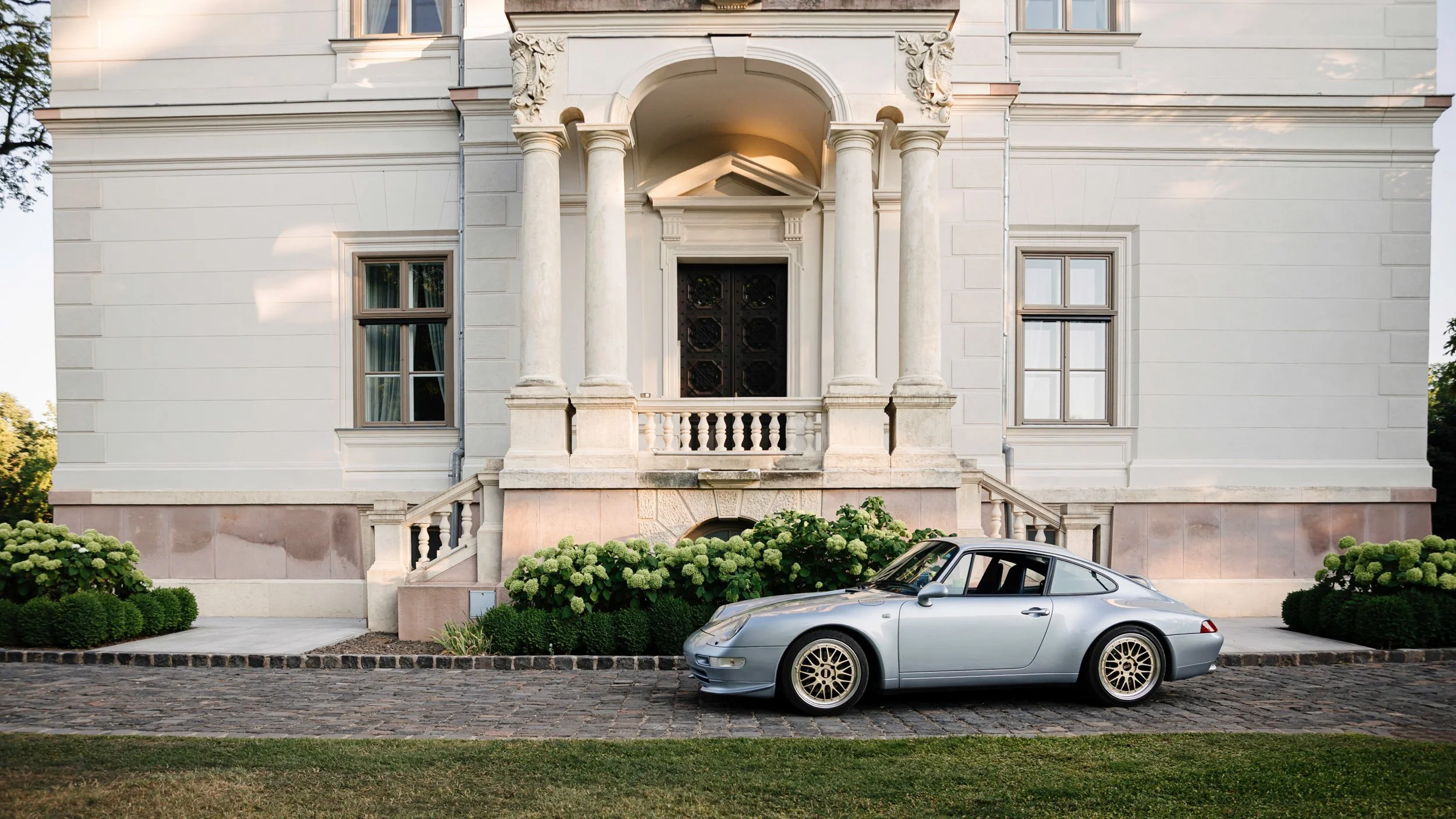 Front view of a grand, white, neoclassical-style house with a portico supported by four columns, ornate decorative details, and large windows. A silver sports car is parked on a cobblestone street in front. There are green bushes and flowers along th