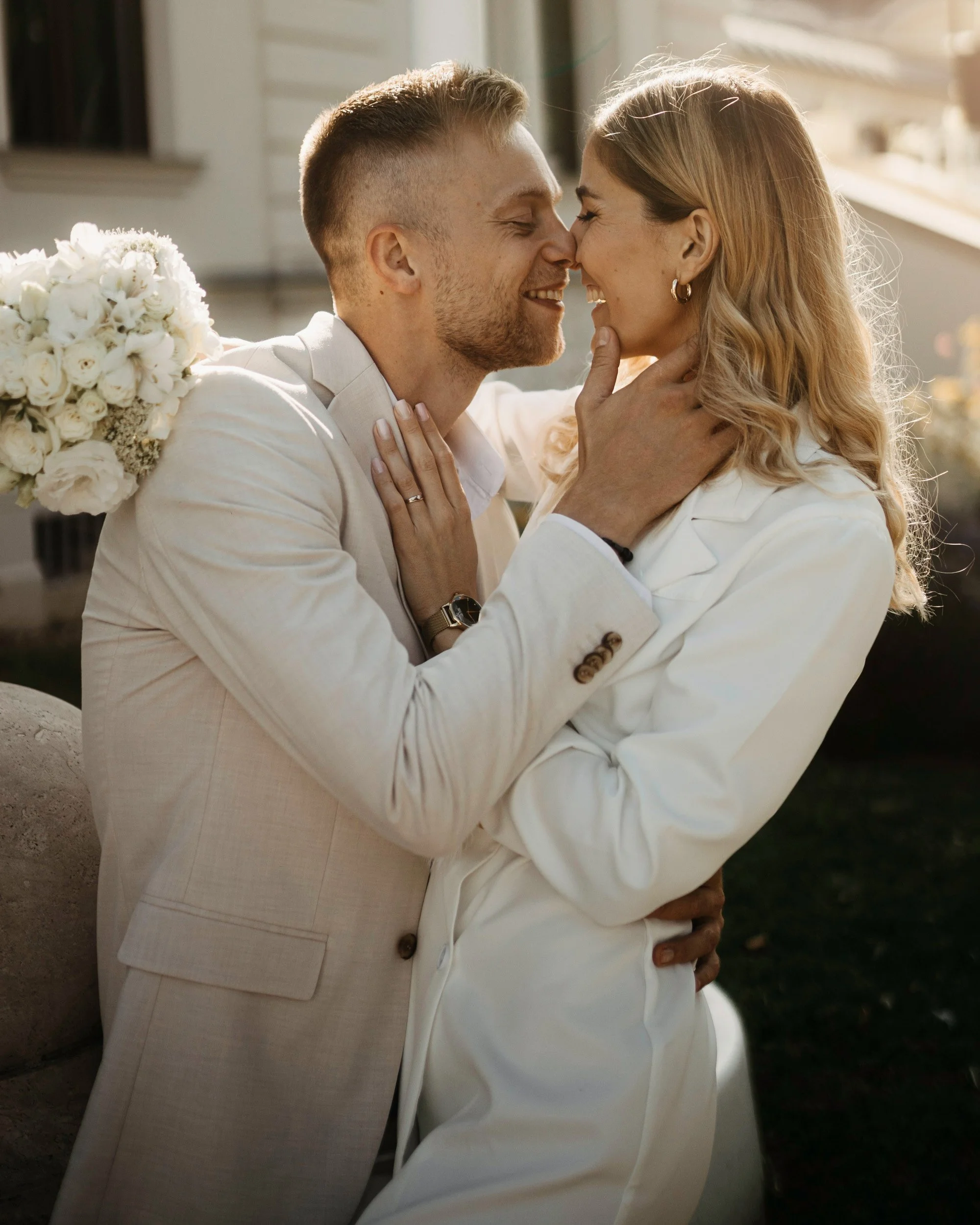 A couple sharing a close, joyful moment with foreheads and noses touching, outdoors, with a woman holding a bouquet of white flowers, both dressed in white.
