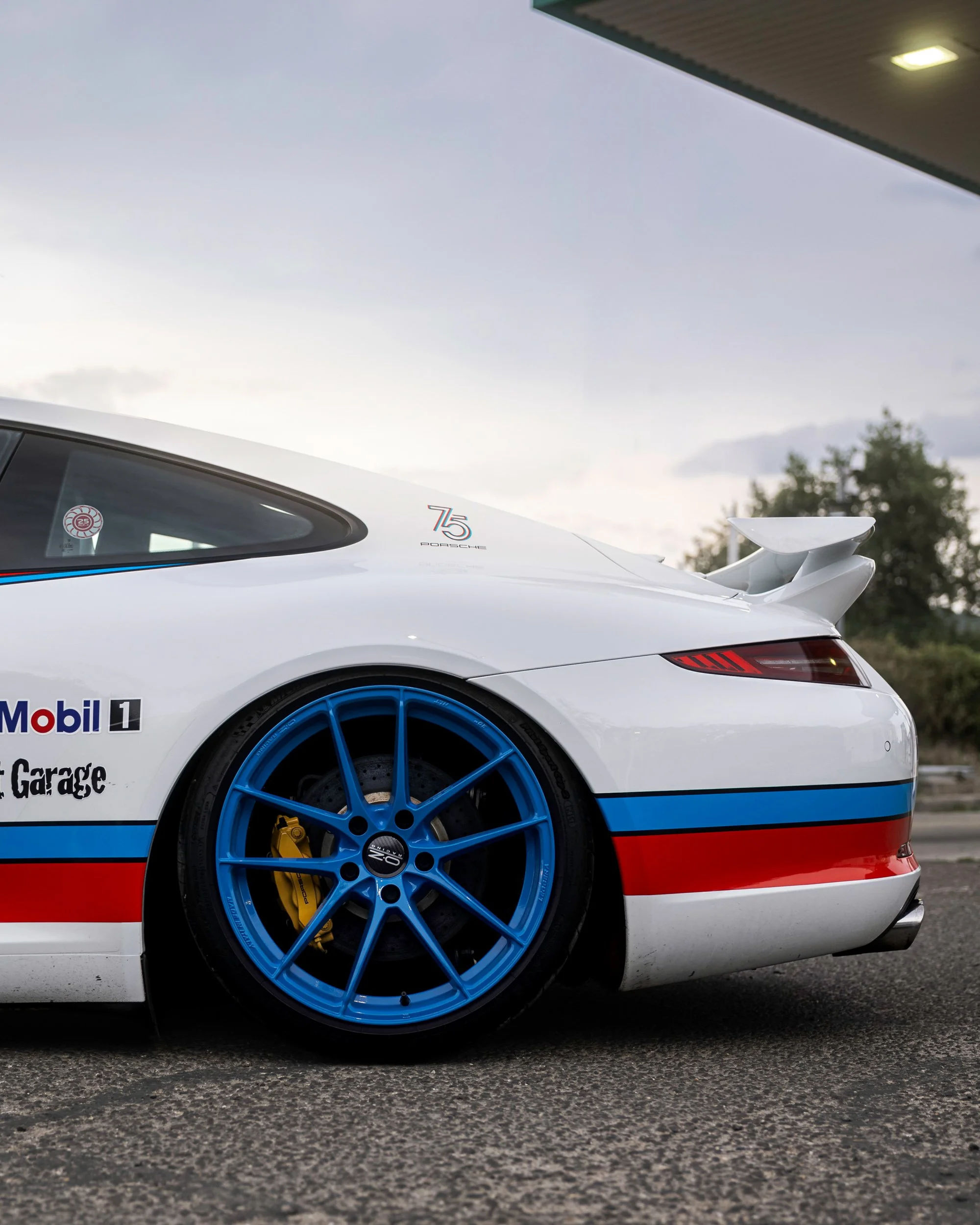 Close-up of a white race car with blue wheels, red accents, and sponsorship decals, including Mobil 1, parked on asphalt, showing part of the rear and side of the vehicle.
