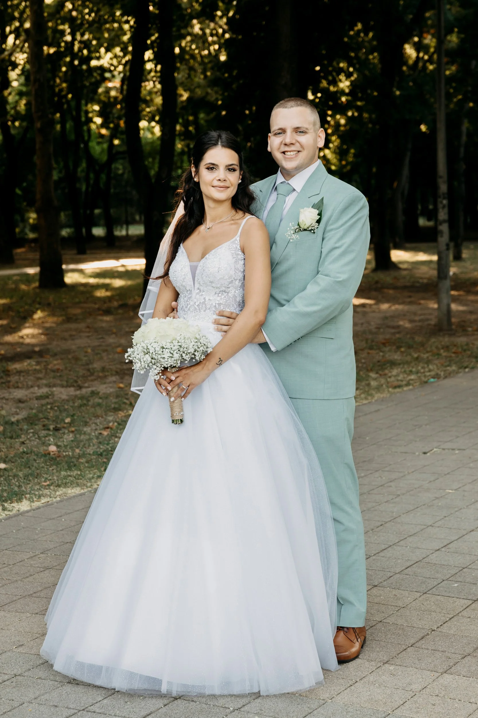 A bride and groom standing together outdoors in a park-like setting with trees in the background. The bride wears a white wedding gown holding a bouquet of white flowers, and the groom is dressed in a light green suit with a white shirt and tie, smil