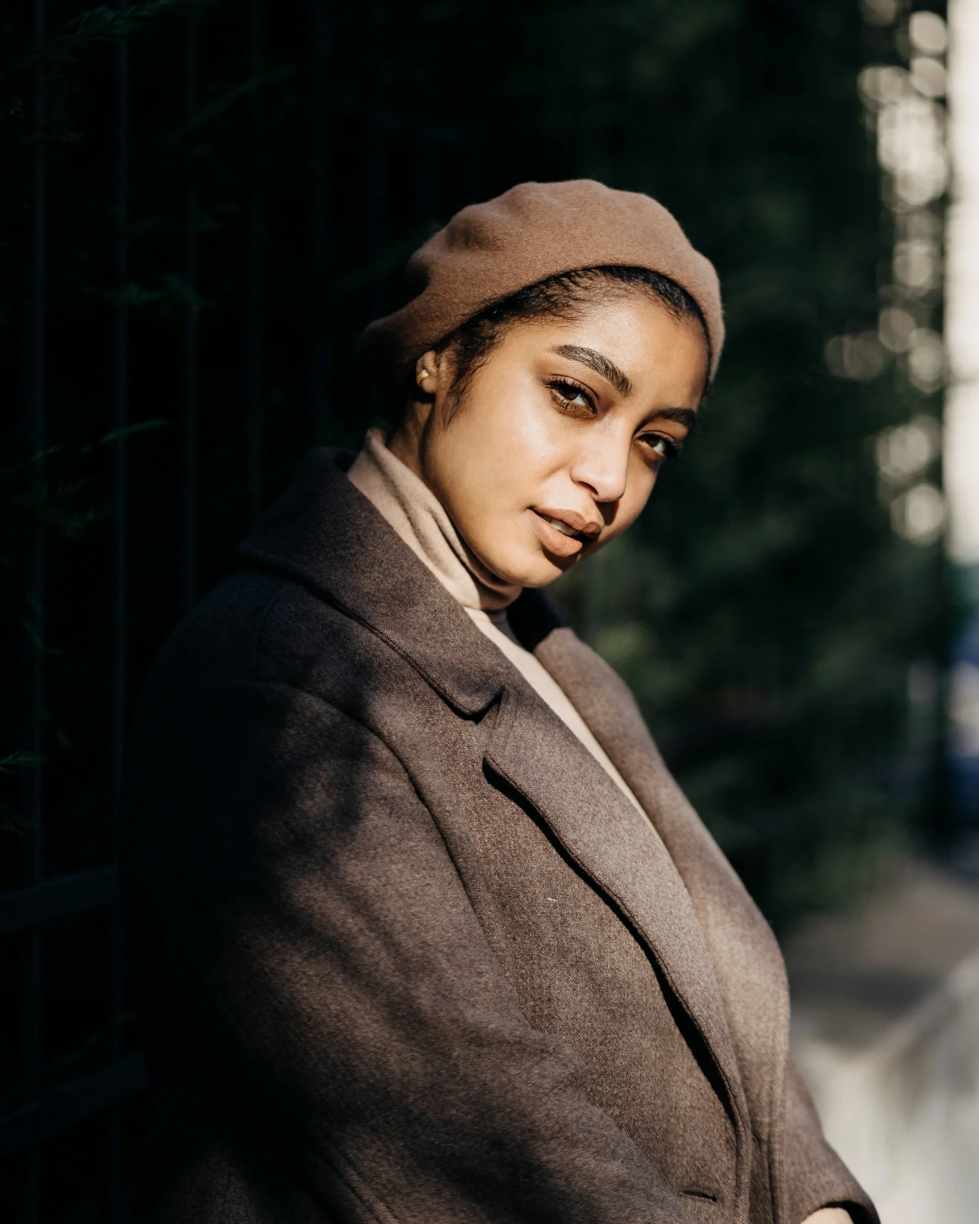 A woman with light brown skin, short curly hair, wearing a tan beret and a dark coat, posing outdoors near a black fence with blurred greenery in the background.