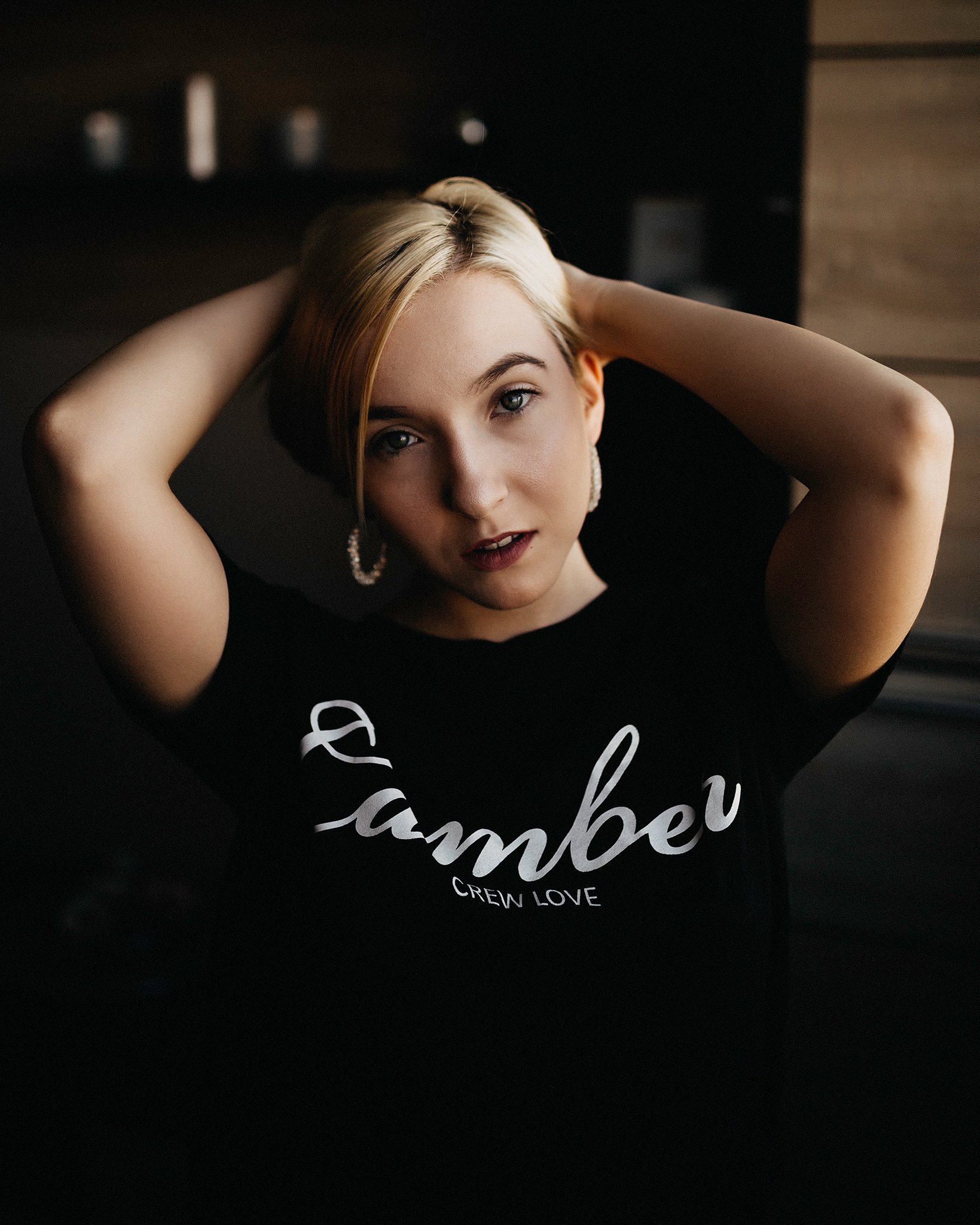 A woman with blonde hair and pearl earrings leaning back with hands behind her head, wearing a black shirt that says 'CREDI LOVE'.