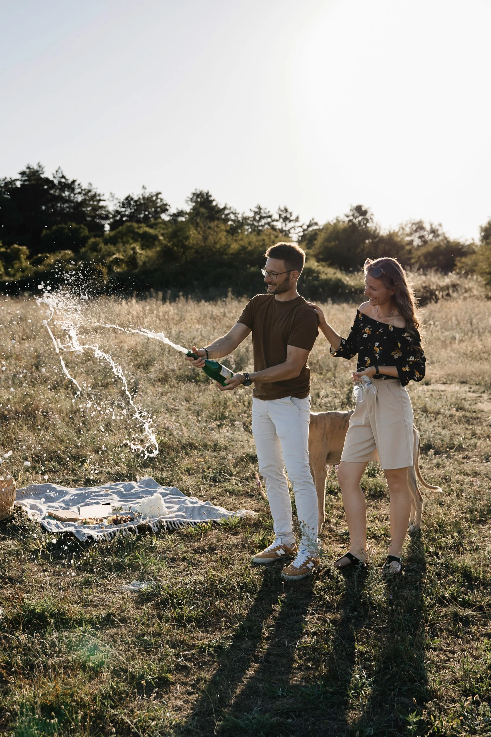 A man and woman holding a champagne bottle and spraying it in a field during sunset, with a dog nearby, and a picnic blanket on the ground.
