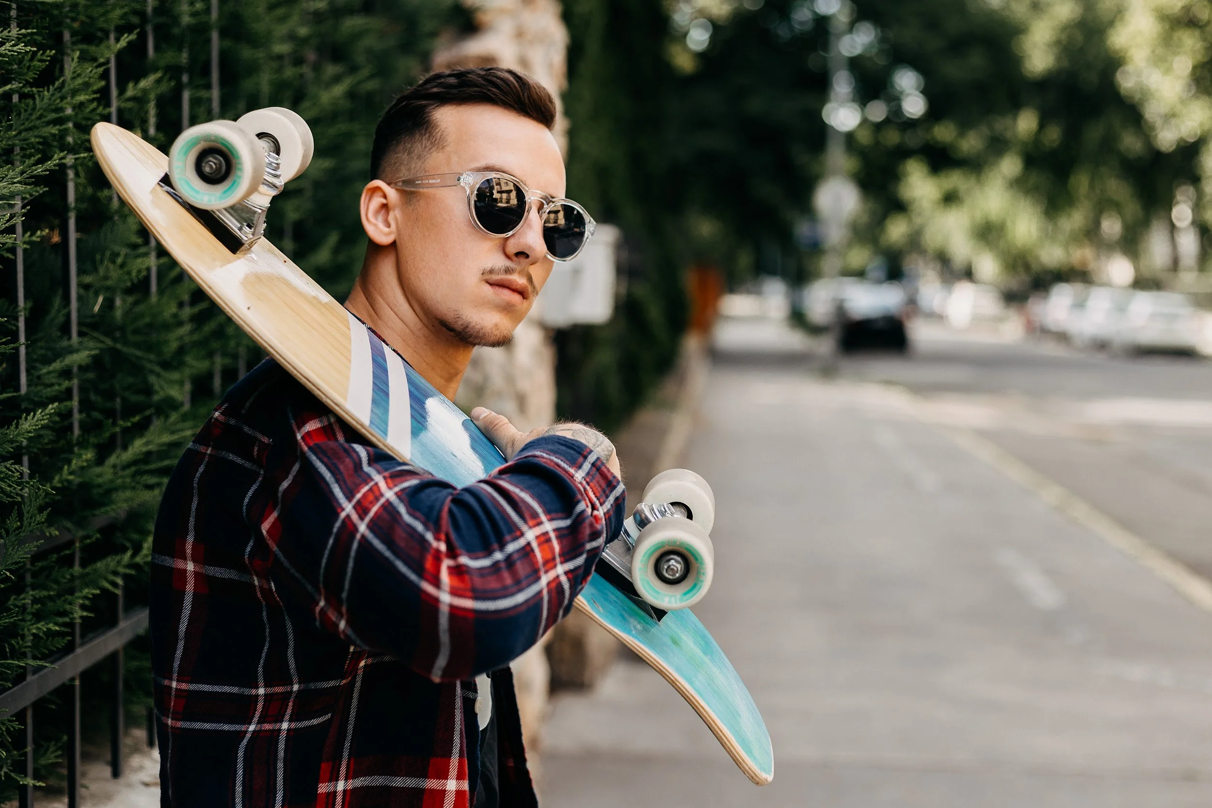 Young man with short brown hair and a goatee, wearing sunglasses and a red and black plaid shirt, holding a skateboard over his shoulder, standing outdoors near a hedge and a blurred street background.