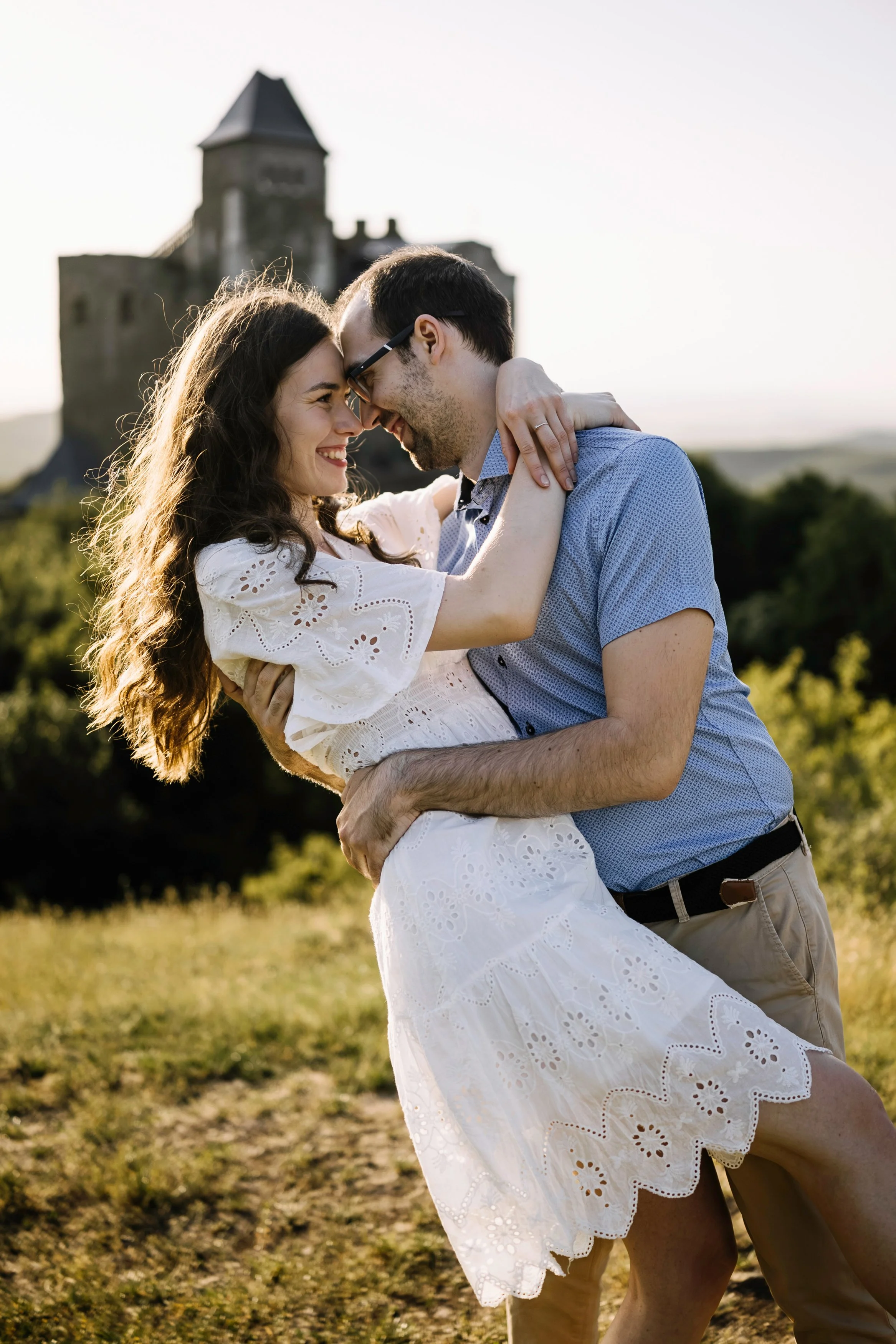 A couple dancing or embracing outdoors near a historic castle or tower during sunset or late afternoon, with green grass and trees in the background.