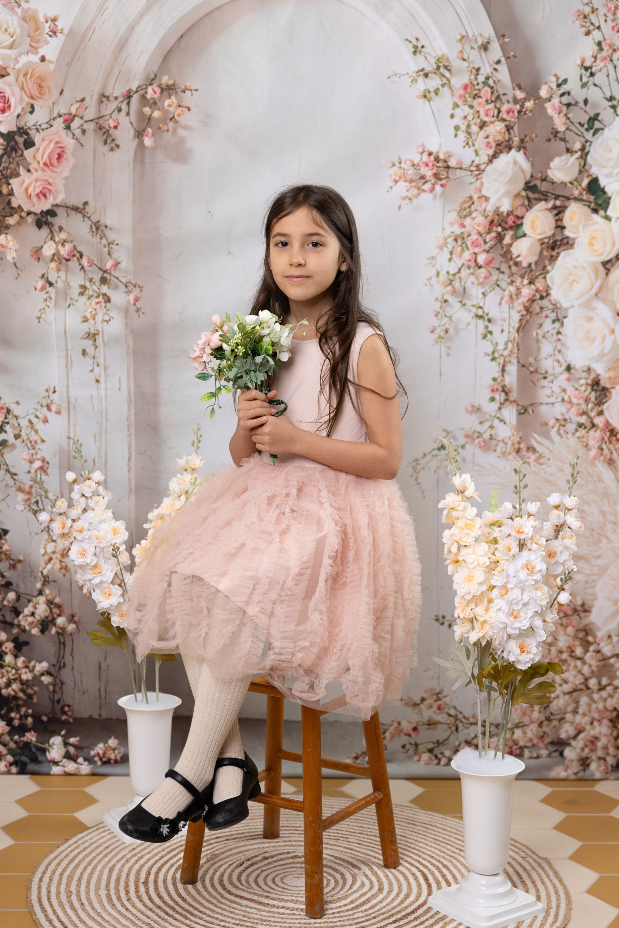 Young girl in a pink dress sitting on a wooden chair, holding a bouquet of flowers, in a floral decorated setting with a patterned rug and two white vases with flowers.