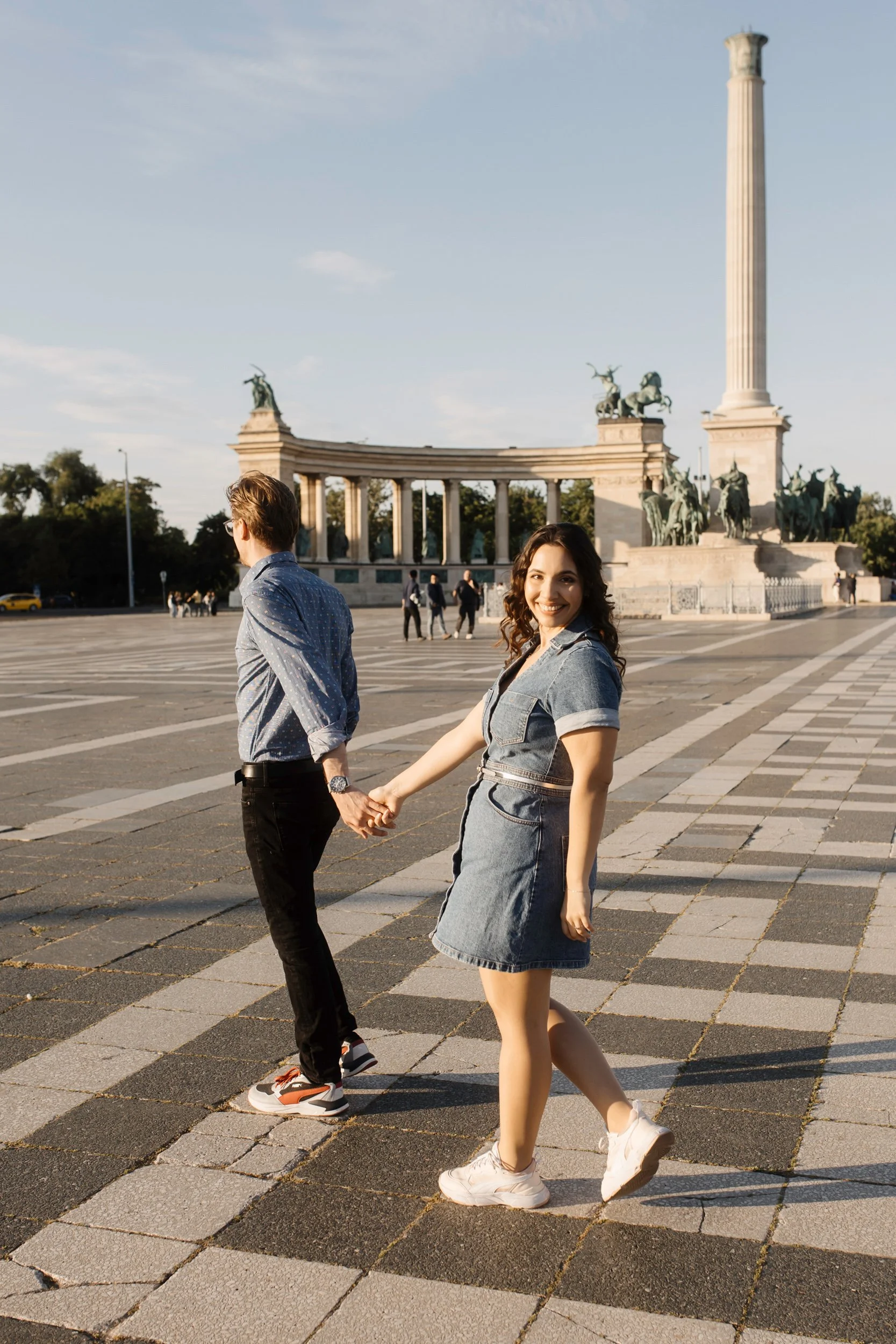 A young woman with curly dark hair smiling and holding hands with a man, walking in front of a monument with statues and a tall column, on a paved square during daytime.