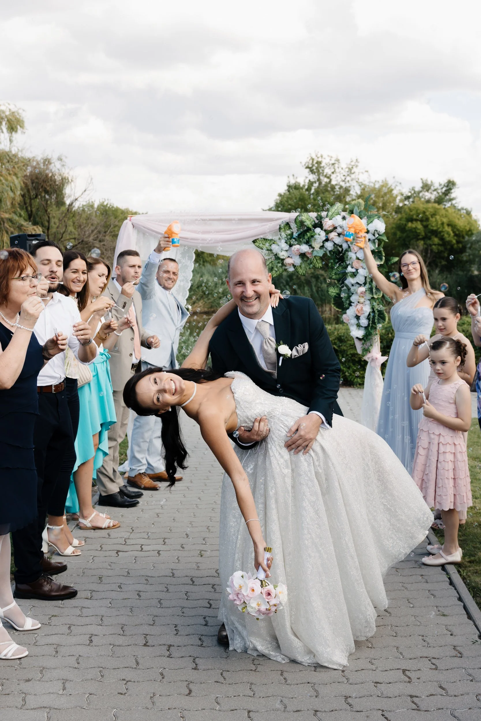 A newlywed couple is seen at their wedding celebration, with the bride in a white wedding dress and the groom in a black suit. The groom is dipping the bride in a dance pose, and they are smiling happily. Guests are surrounding them, blowing bubbles,