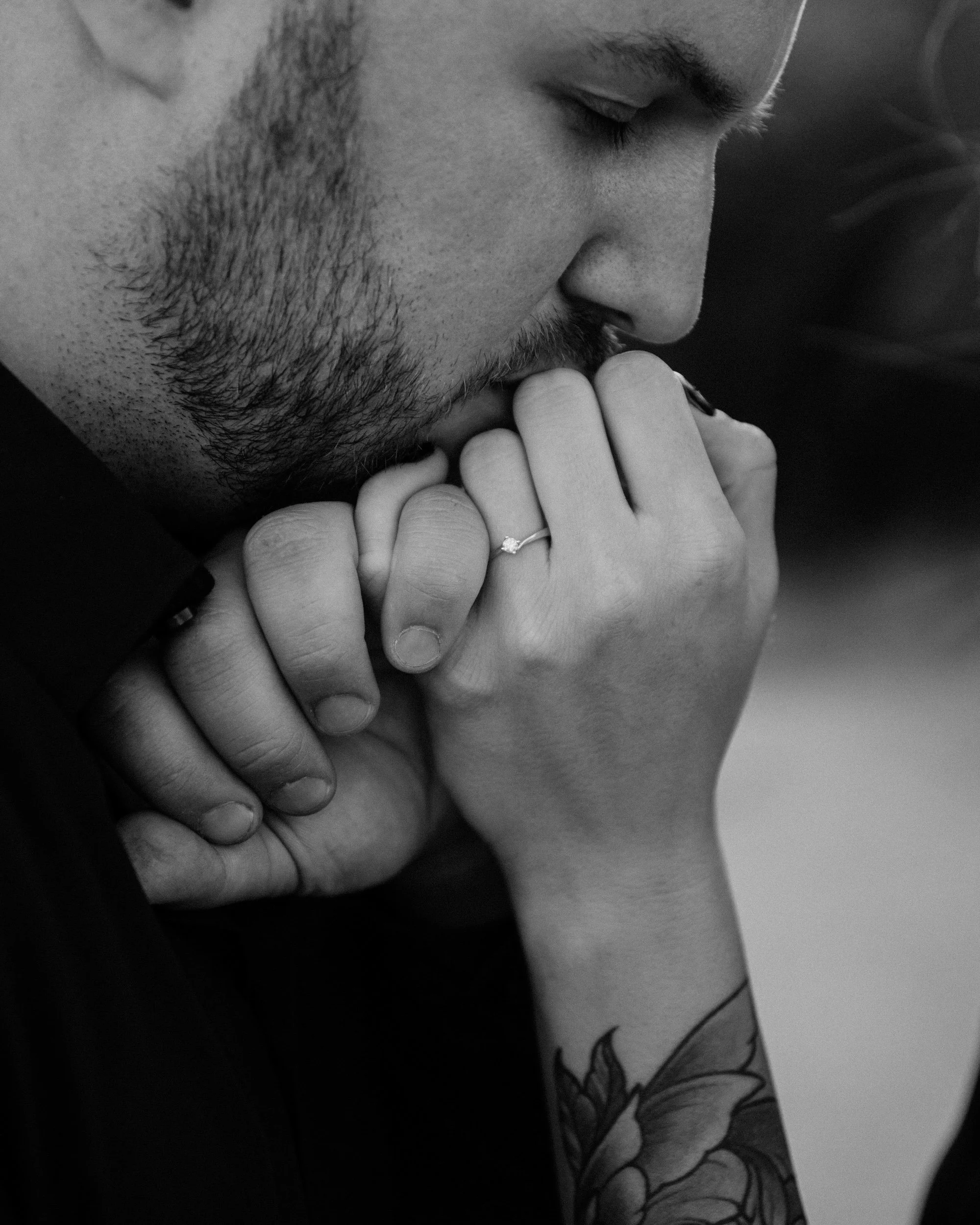 Close-up black-and-white photo of a man with a beard and closed eyes, holding a woman's hand with an engagement ring near his lips. Part of a tattoo is visible on the woman's arm.
