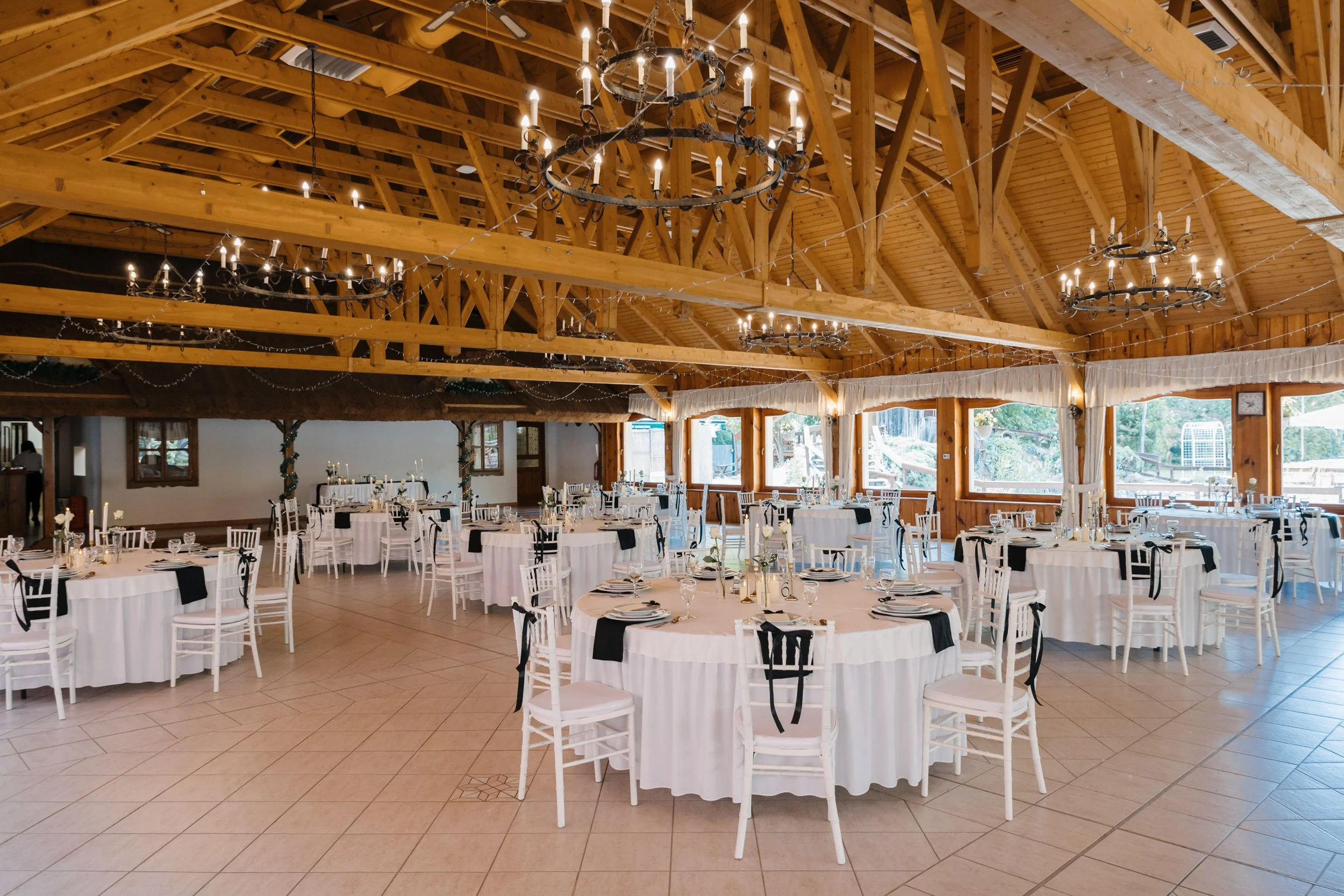 Wedding reception hall decorated with round tables covered in white tablecloths, black and white chair decorations, tall white candles in glass holders, and elegant place settings. The hall has wooden beams and chandeliers hanging from the ceiling, w