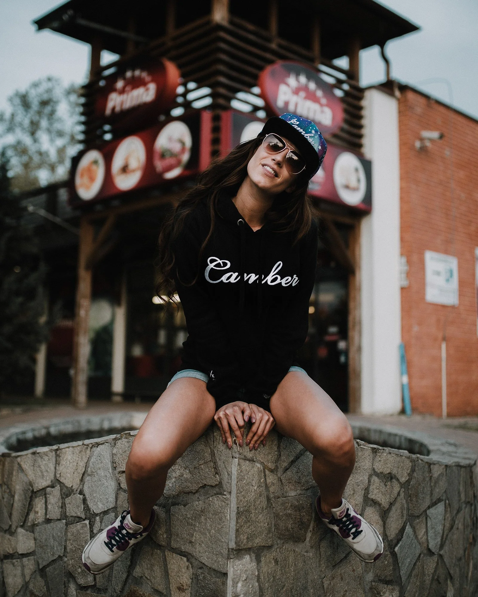 A woman wearing sunglasses, a cap, a black hoodie, and sneakers sitting on a stone ledge with her legs spread apart in front of a building with signs that say "Chamber" and "Famous".
