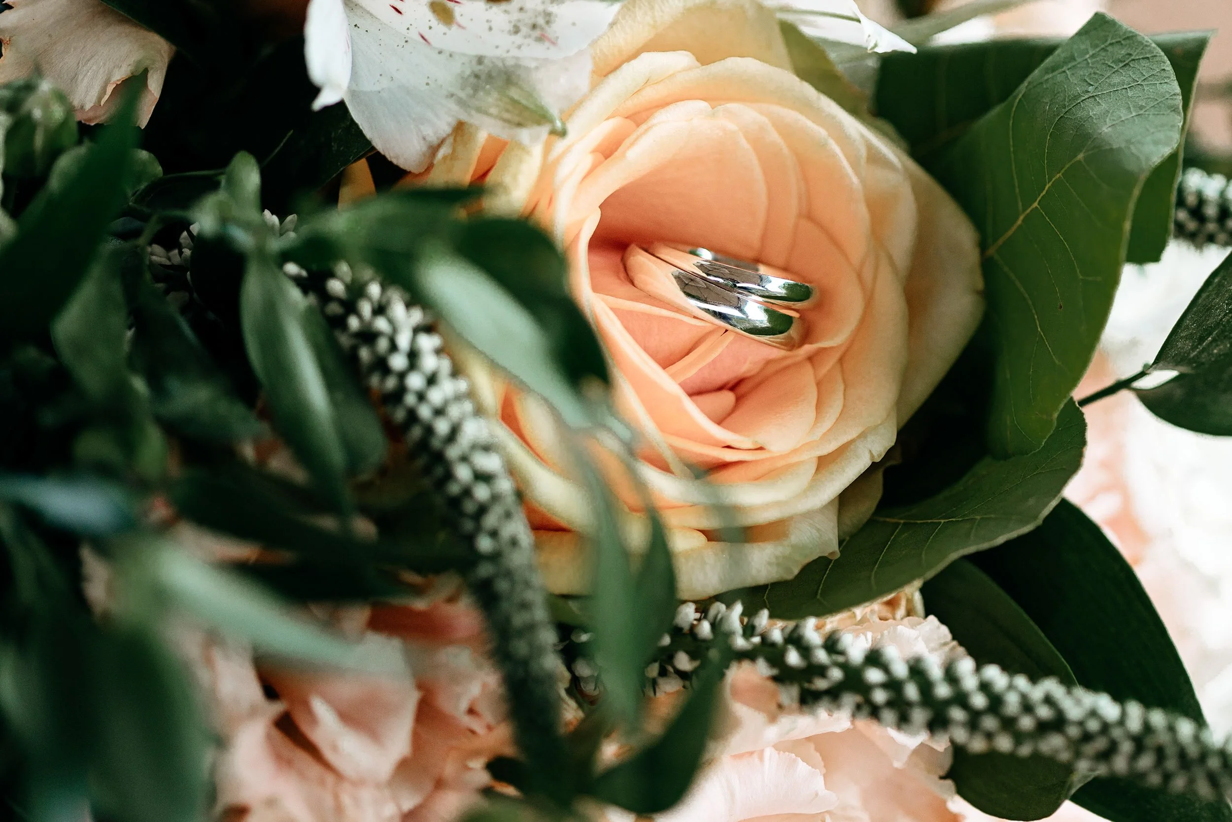 A close-up of a wedding or engagement ring nestled inside a light peach-colored rose, surrounded by green foliage and smaller white flowers, suggesting a floral arrangement or bouquet.