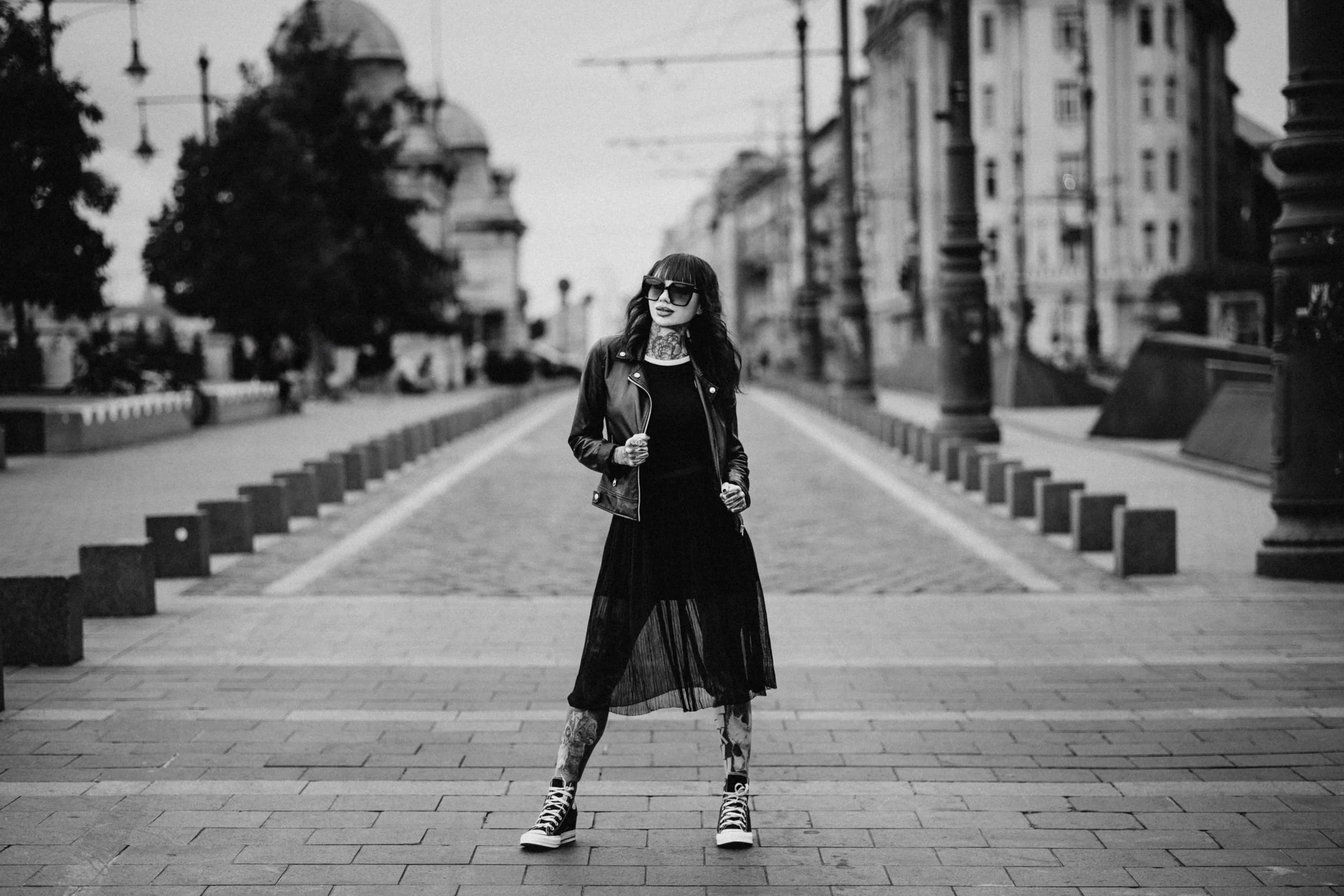 A woman with tattoos on her neck and face, wearing sunglasses, a leather jacket, a long skirt, and high-top sneakers, stands in the middle of a city street in black and white.