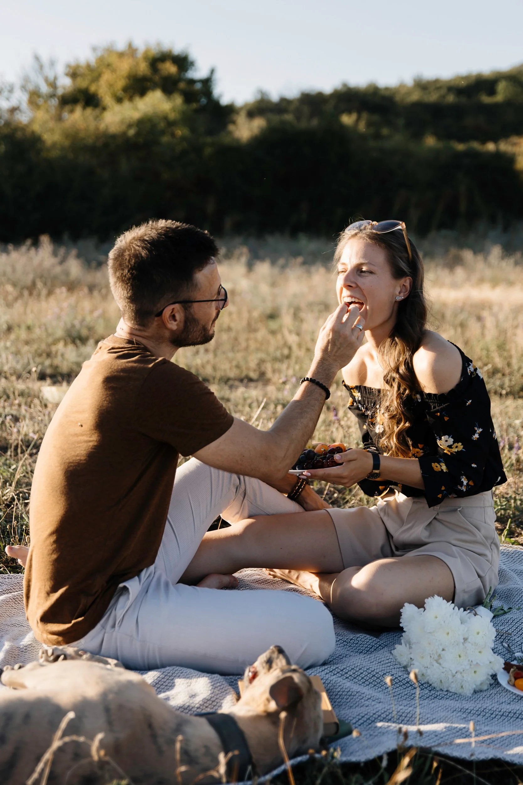 A man and woman sitting on a blanket outdoors, sharing a moment of affection, with dogs lying nearby in a natural, grassy setting during the daytime.
