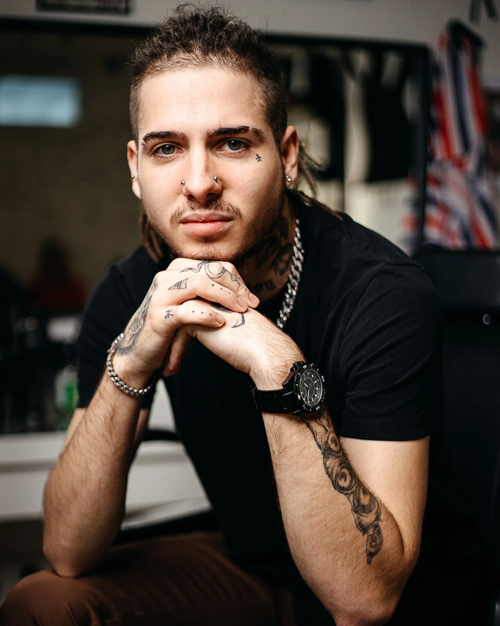 Close-up of a young man with tattoos on his arm and face, wearing a black shirt, silver jewelry, and a watch, sitting indoors with a confident expression.